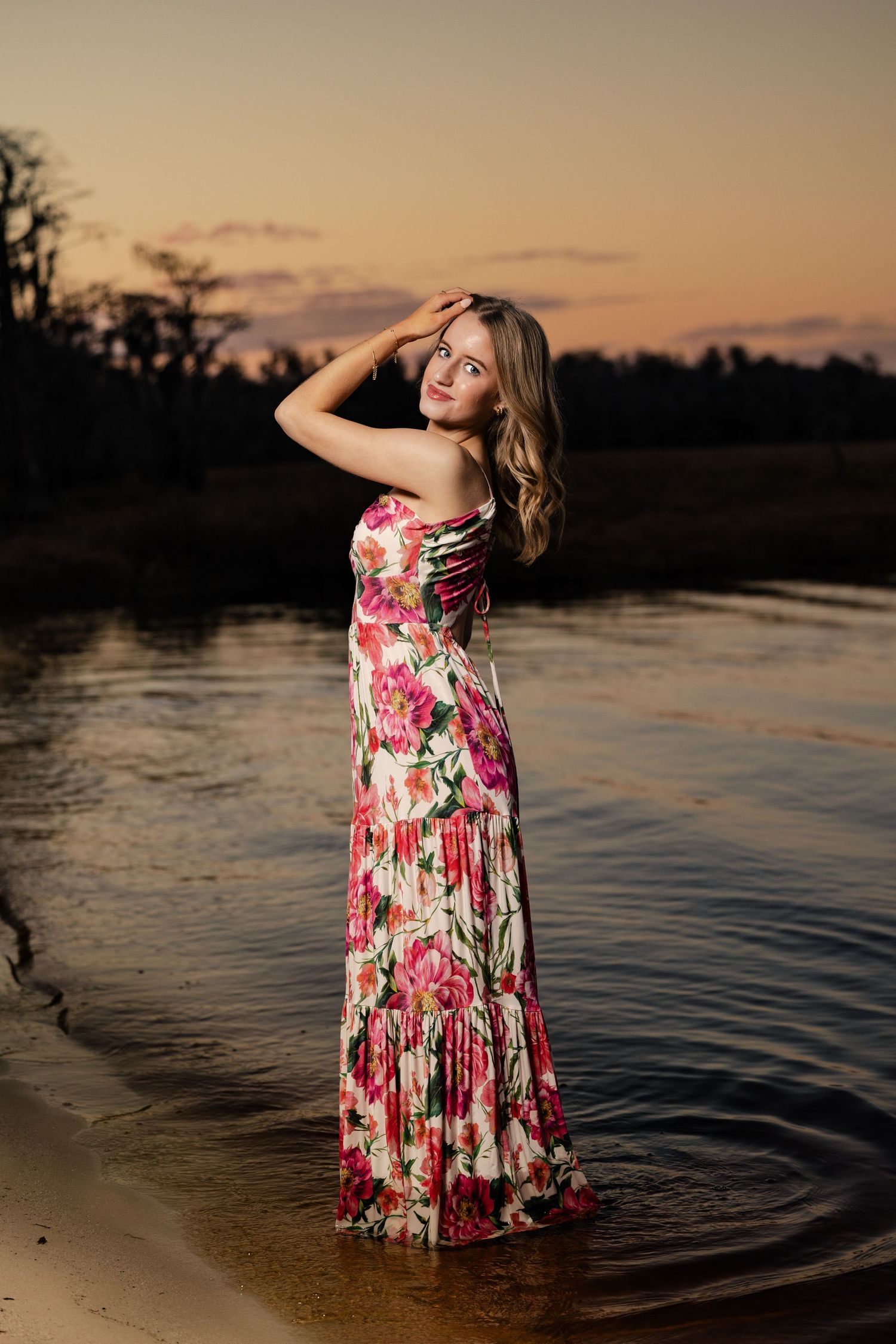 Blonde woman in long floral dress standing in shallow water at sunset.