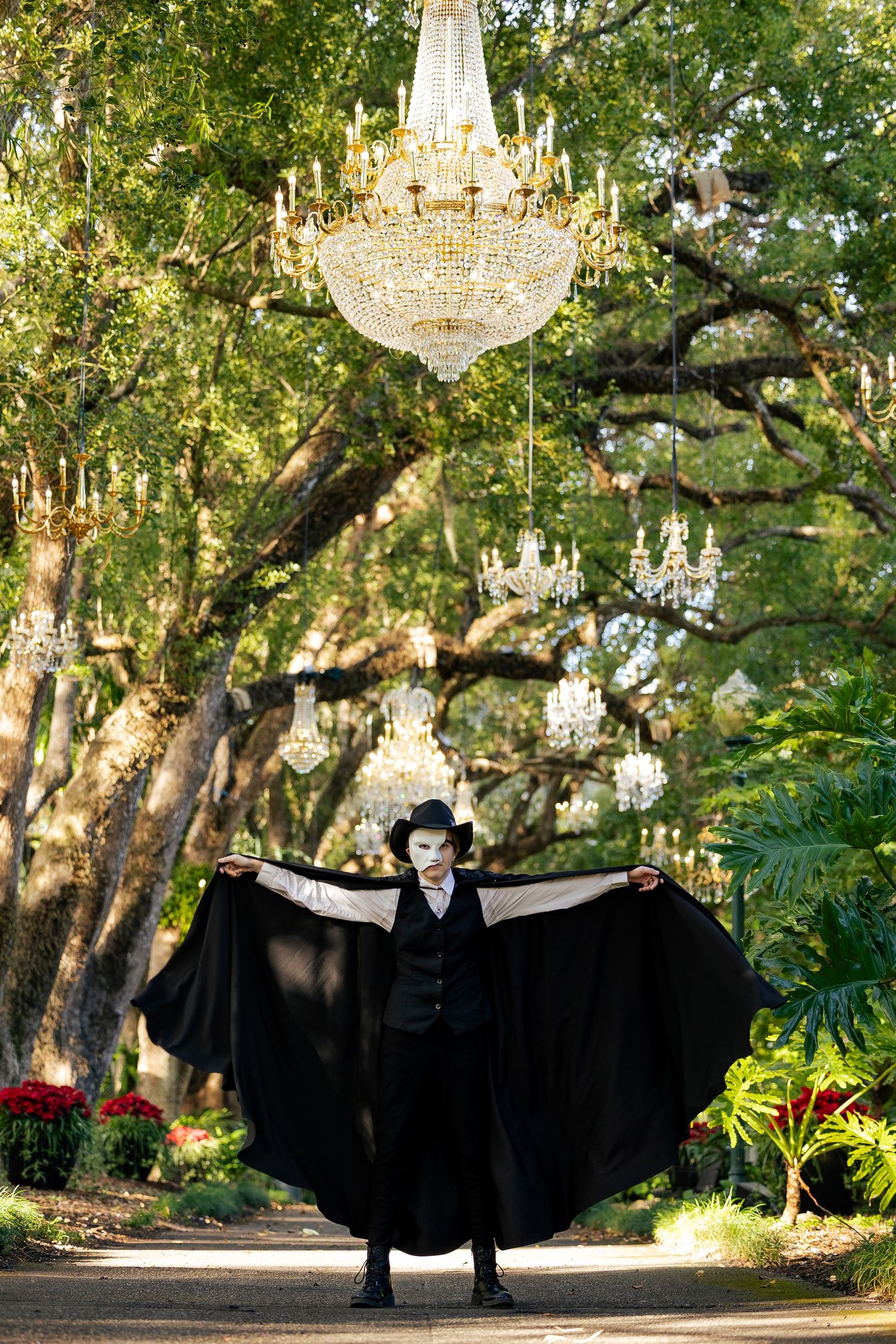 Mysterious masked performer in black cape spreading wings under grand crystal chandelier in lush garden setting.