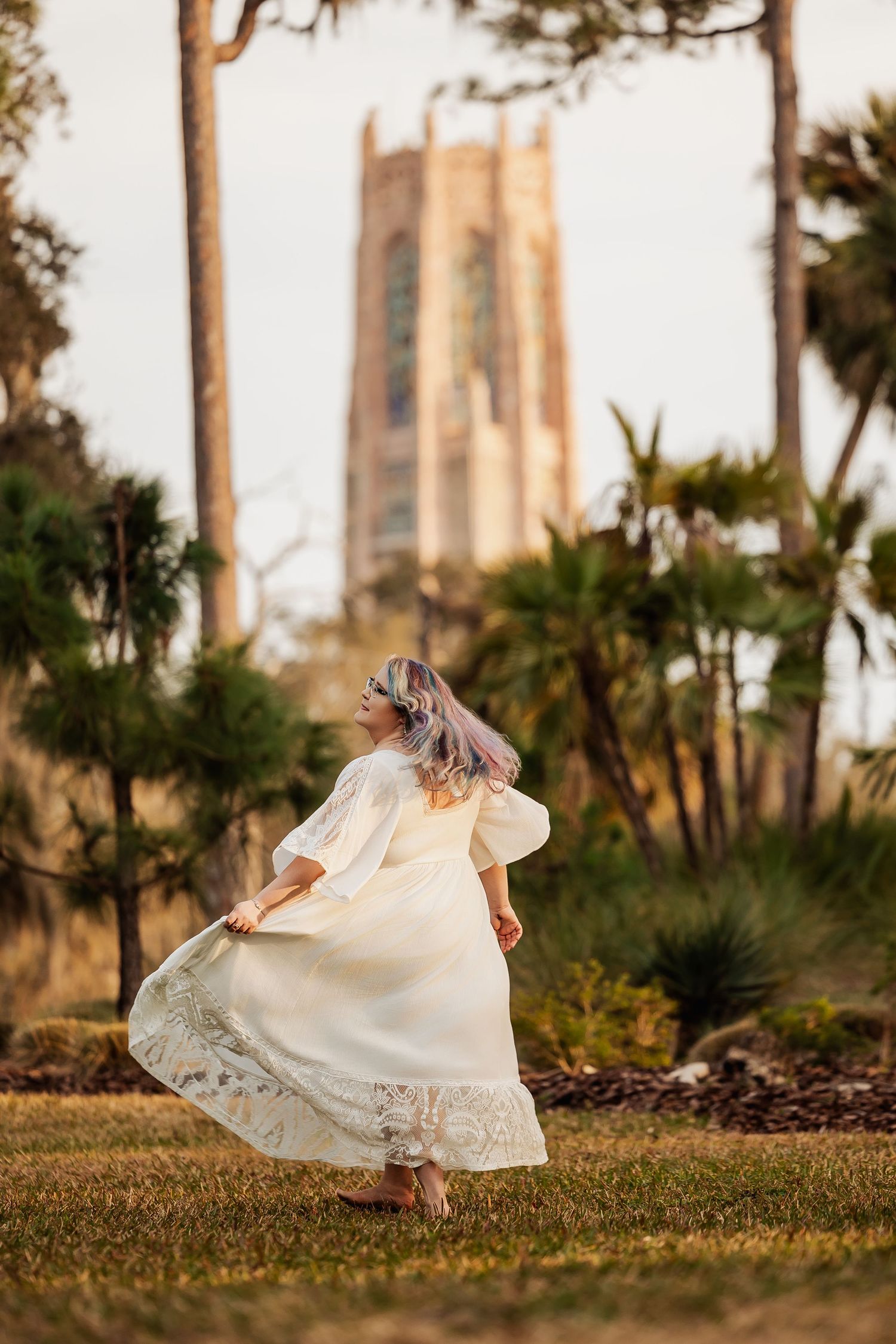 A young girl in a white lace dress twirls in a garden with a tall stone tower and palm trees in the background.
