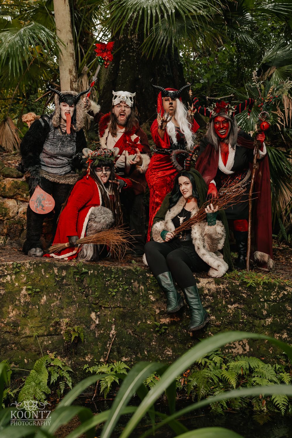 Group of seven people in elaborate fantasy devil and creature costumes pose in lush tropical garden.