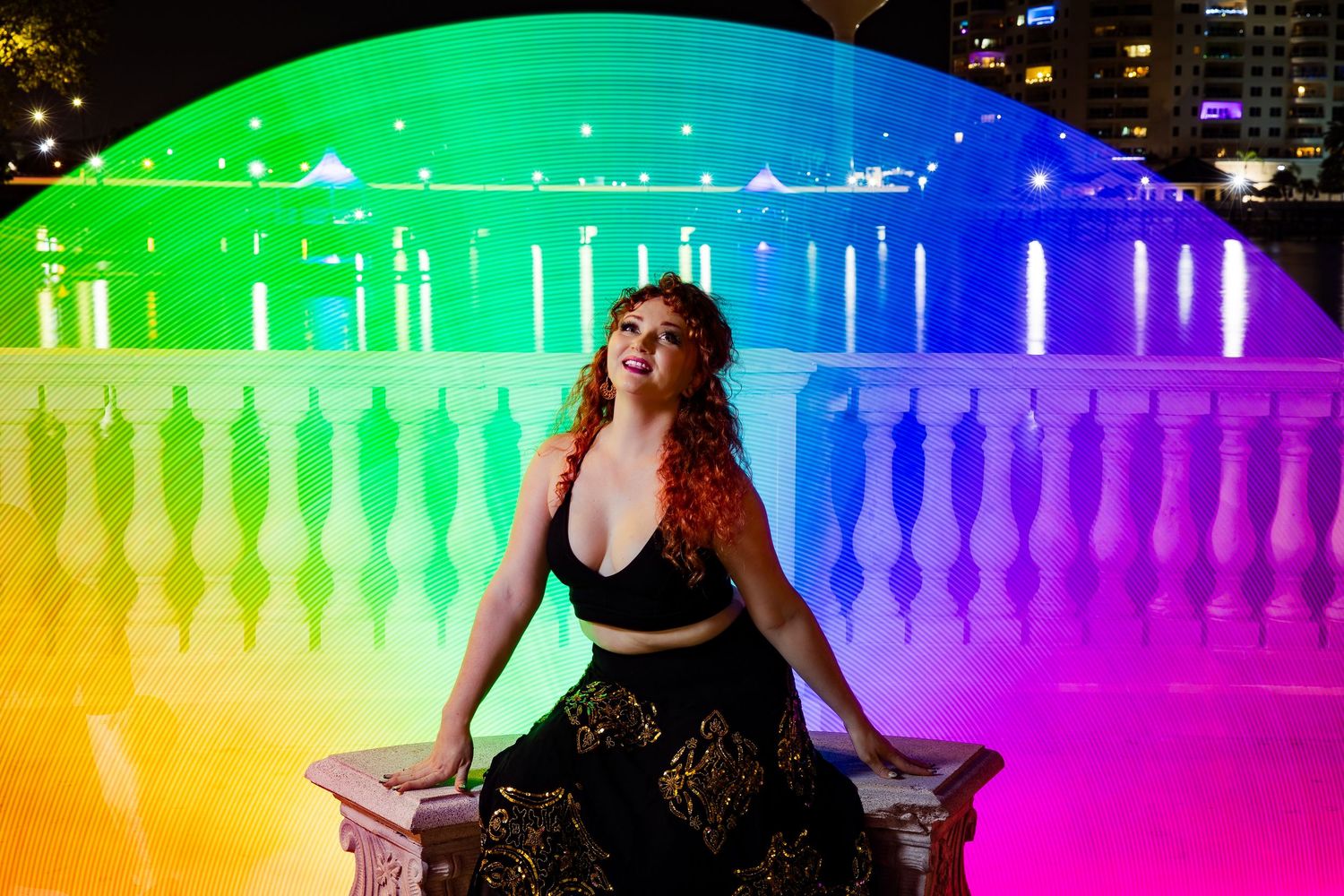 Woman in black dress poses on bench bathed in vibrant rainbow light by a waterfront.