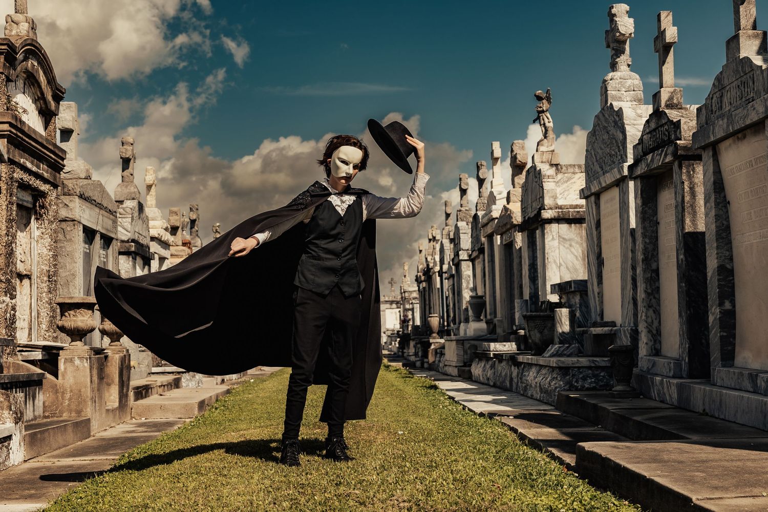 Masked phantom in flowing black cape walks dramatically among white stone tombs in a cemetery.