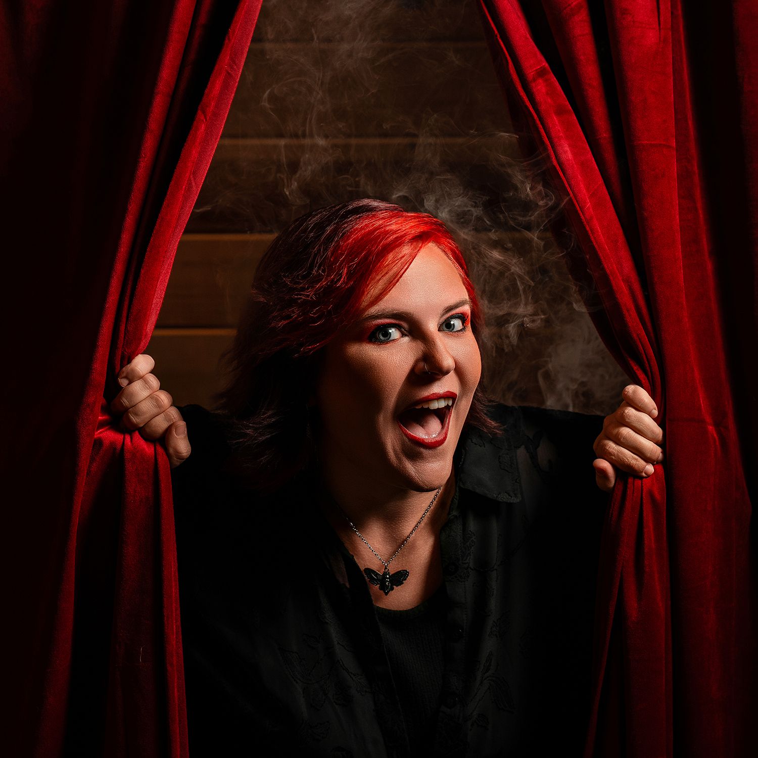 Woman with red hair parting red curtains dramatically, wearing black clothing and necklace, mouth open in excitement.