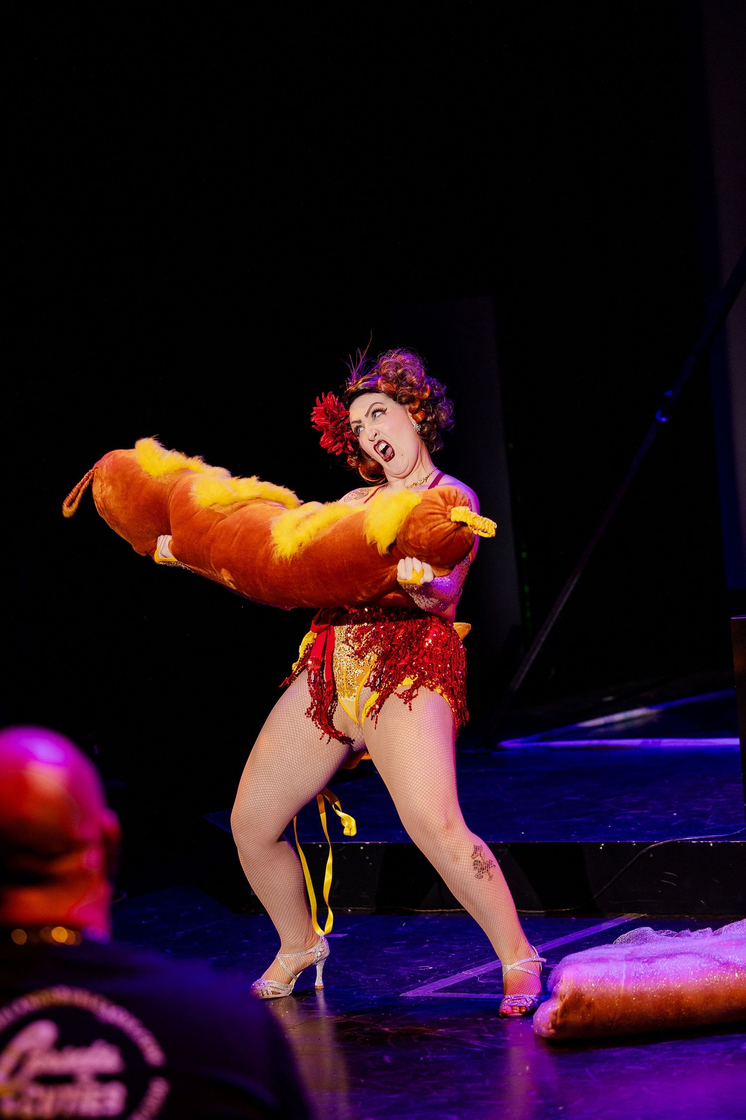 A burlesque performer in a red sequin costume holds a giant hot dog prop on stage during a cabaret performance.