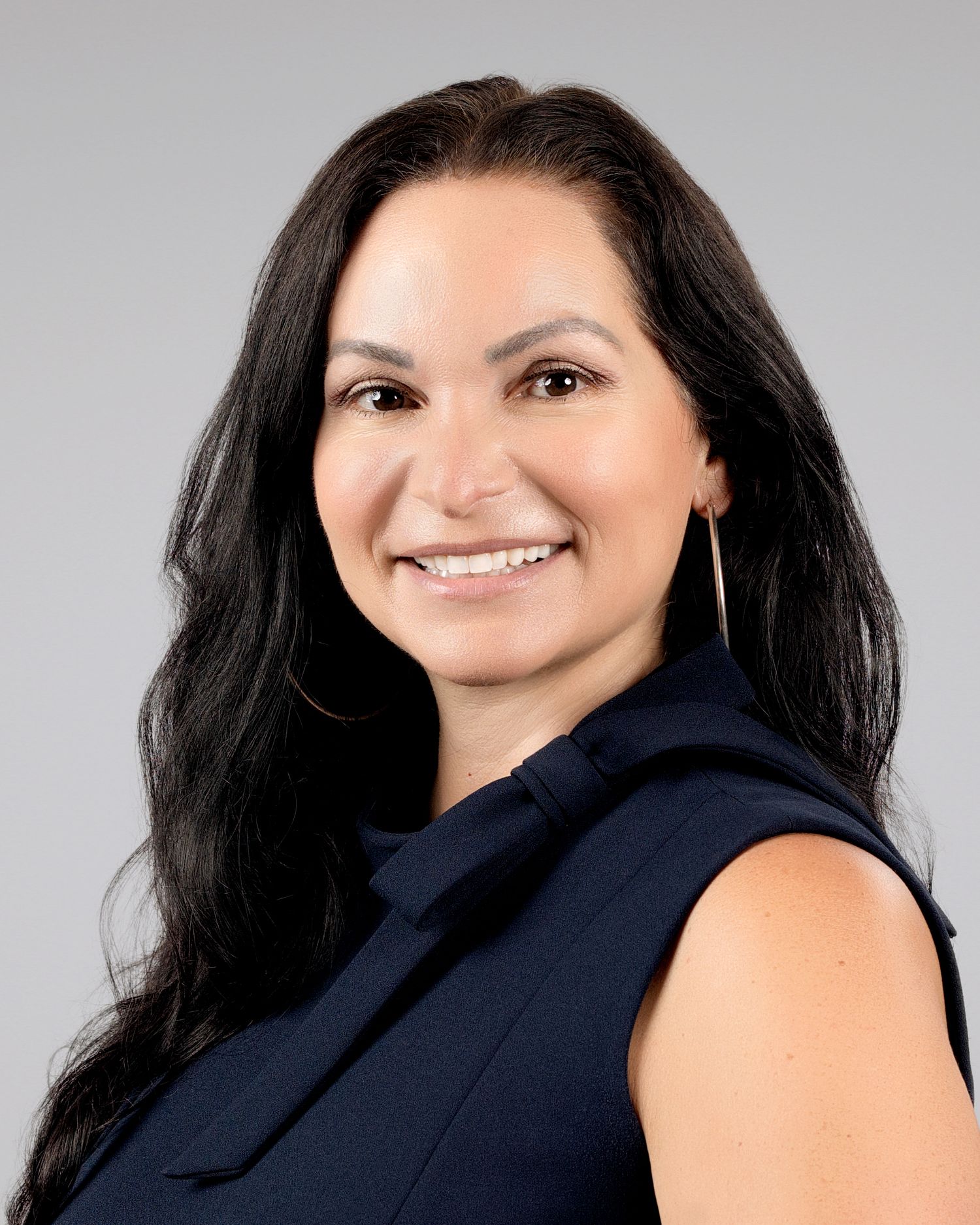 Confident woman with long dark hair wearing a navy sleeveless top poses for a professional business headshot.