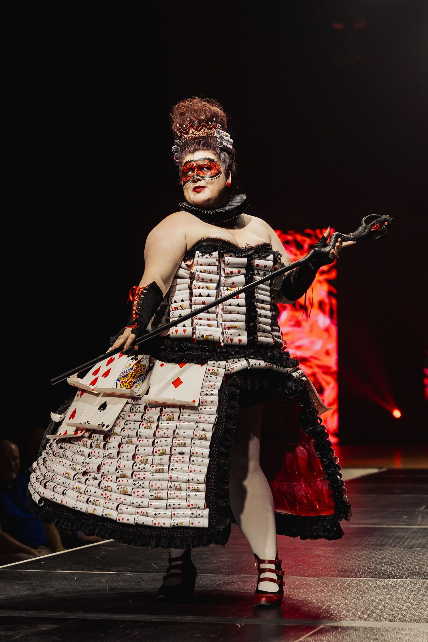 Model in playing card corset dress with mask and scepter poses on dark fashion runway.
