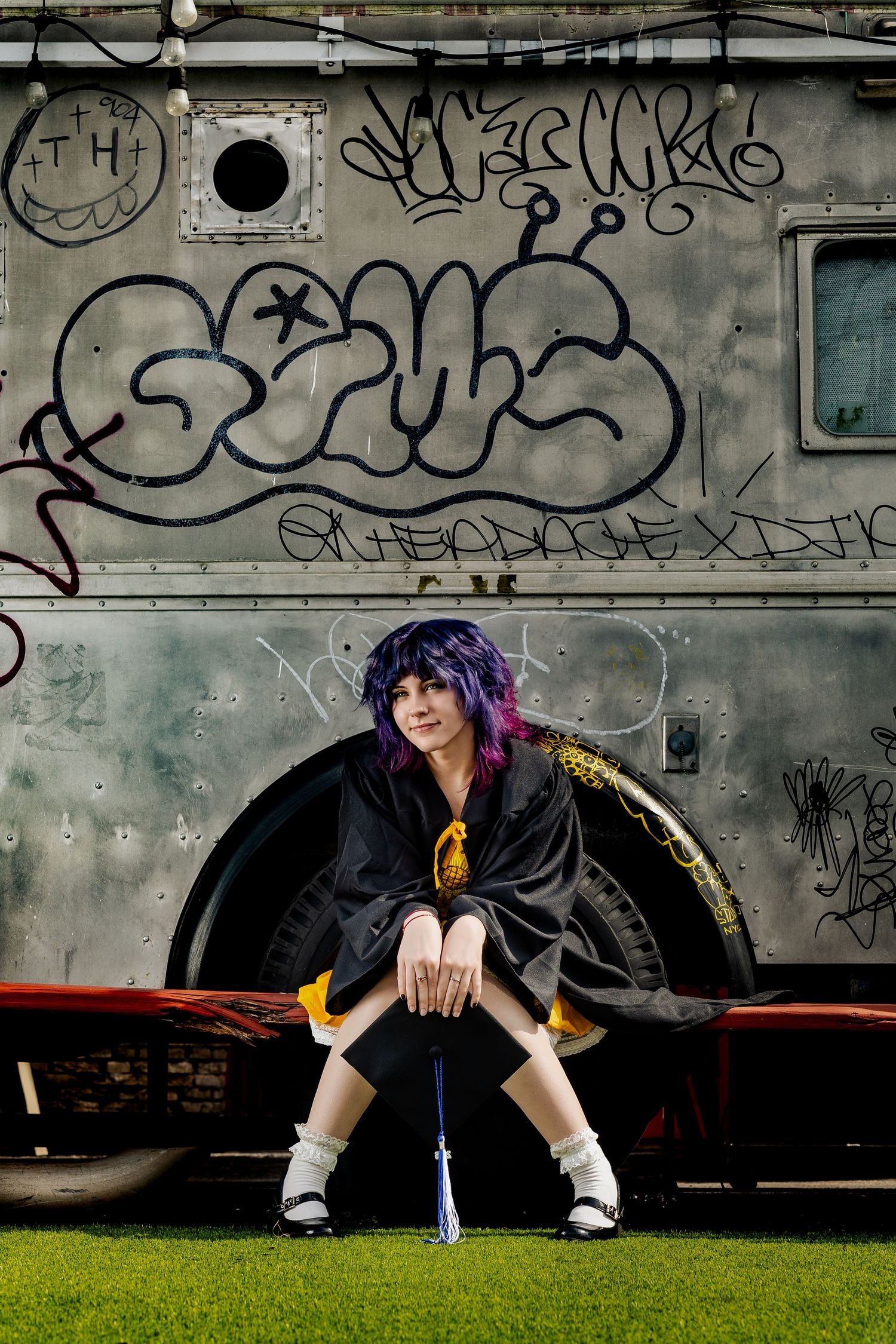 Purple-haired graduate in black cap and gown sits before graffiti-covered vehicle on green turf.