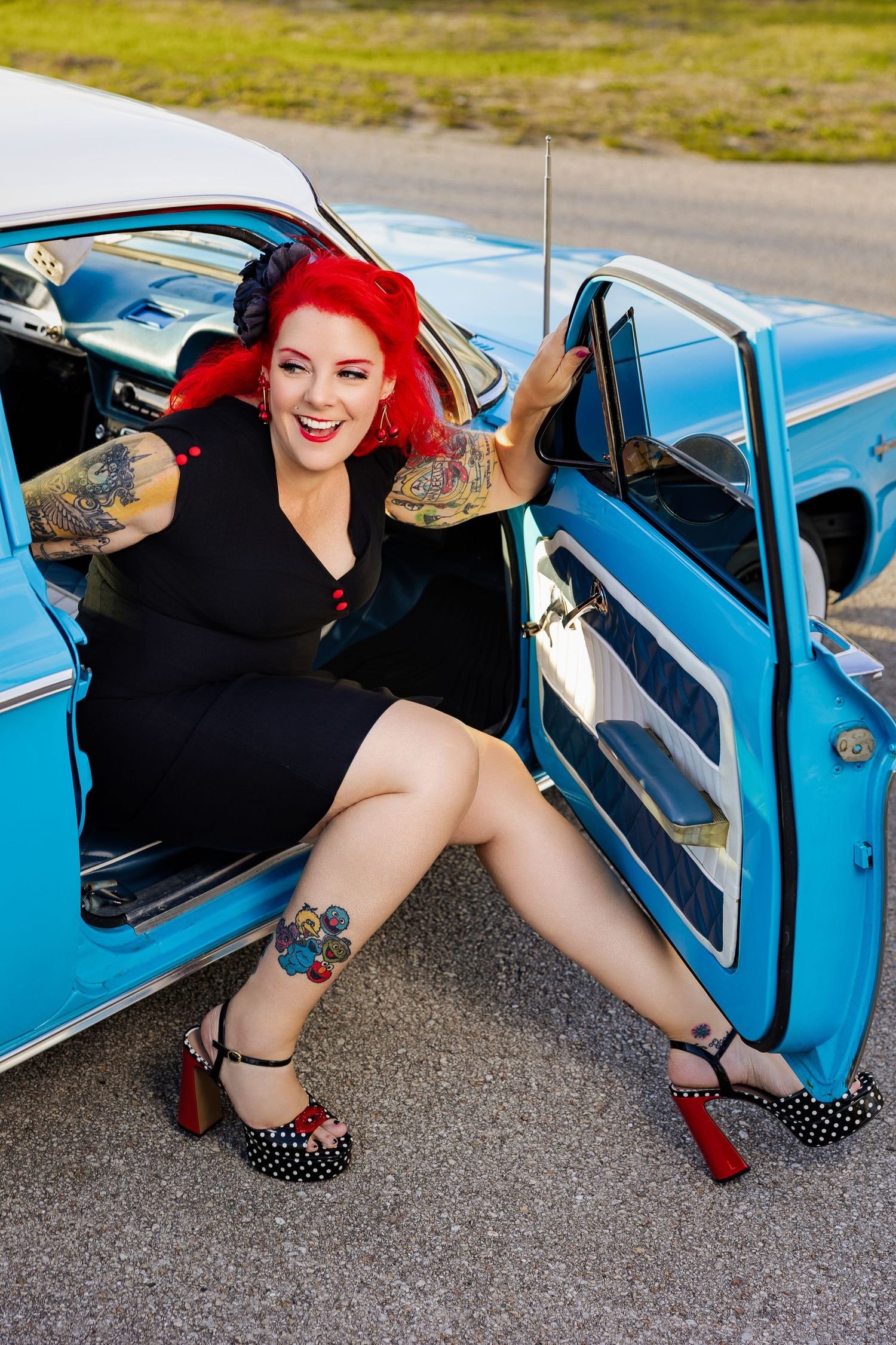 Tattooed redhead pinup woman sitting in open door of blue vintage classic car.