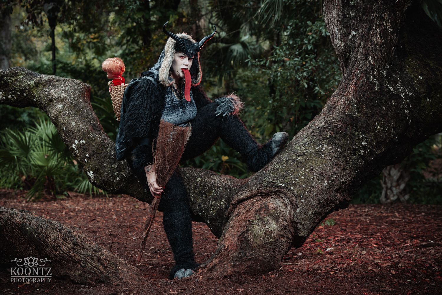 Person in dark horned creature costume perches on a tree branch in a moody forest setting.