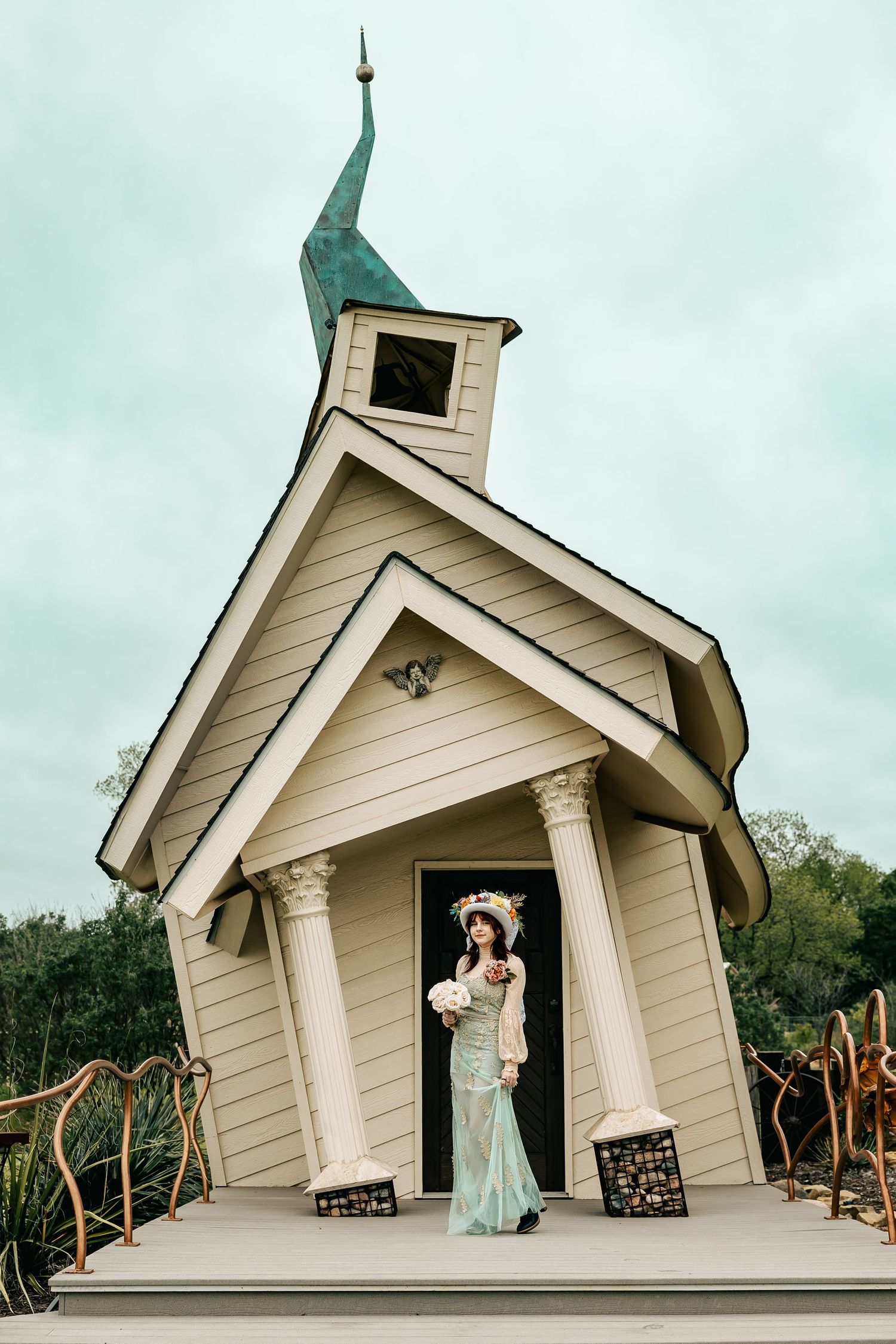 Bride in mint lace gown and floral hat holds bouquet outside charming white chapel with green spire.