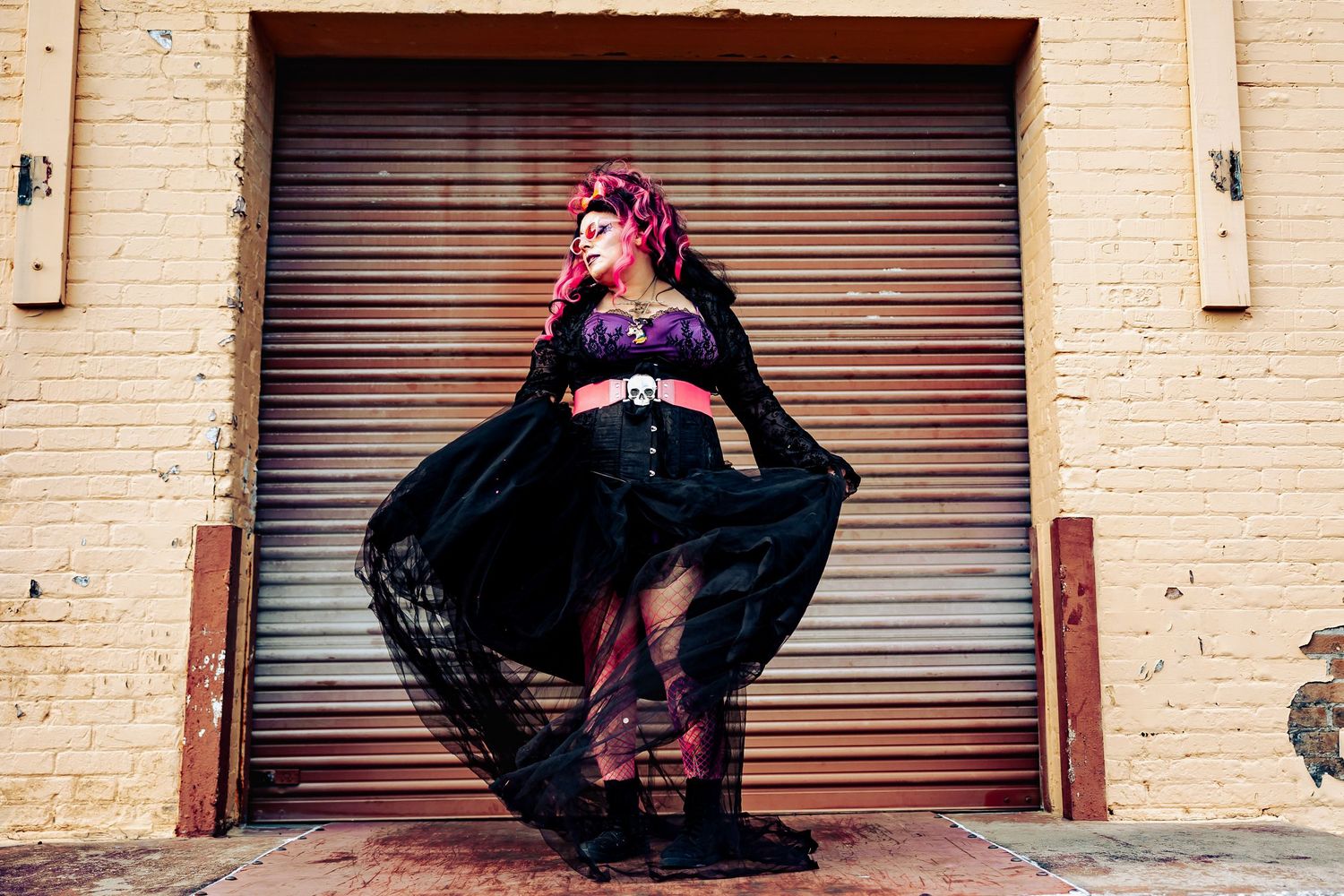 Woman in black lace cape and purple corset stands dramatically before a metal garage door.