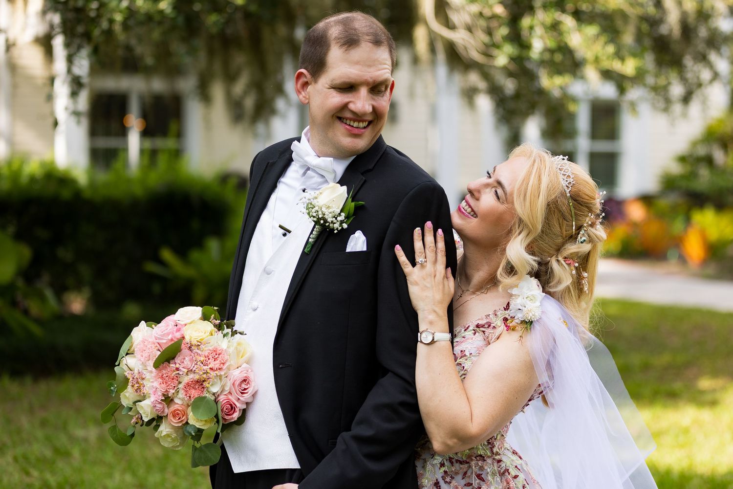 Smiling bride showing wedding ring while groom holds pink rose bouquet in sunny garden setting.