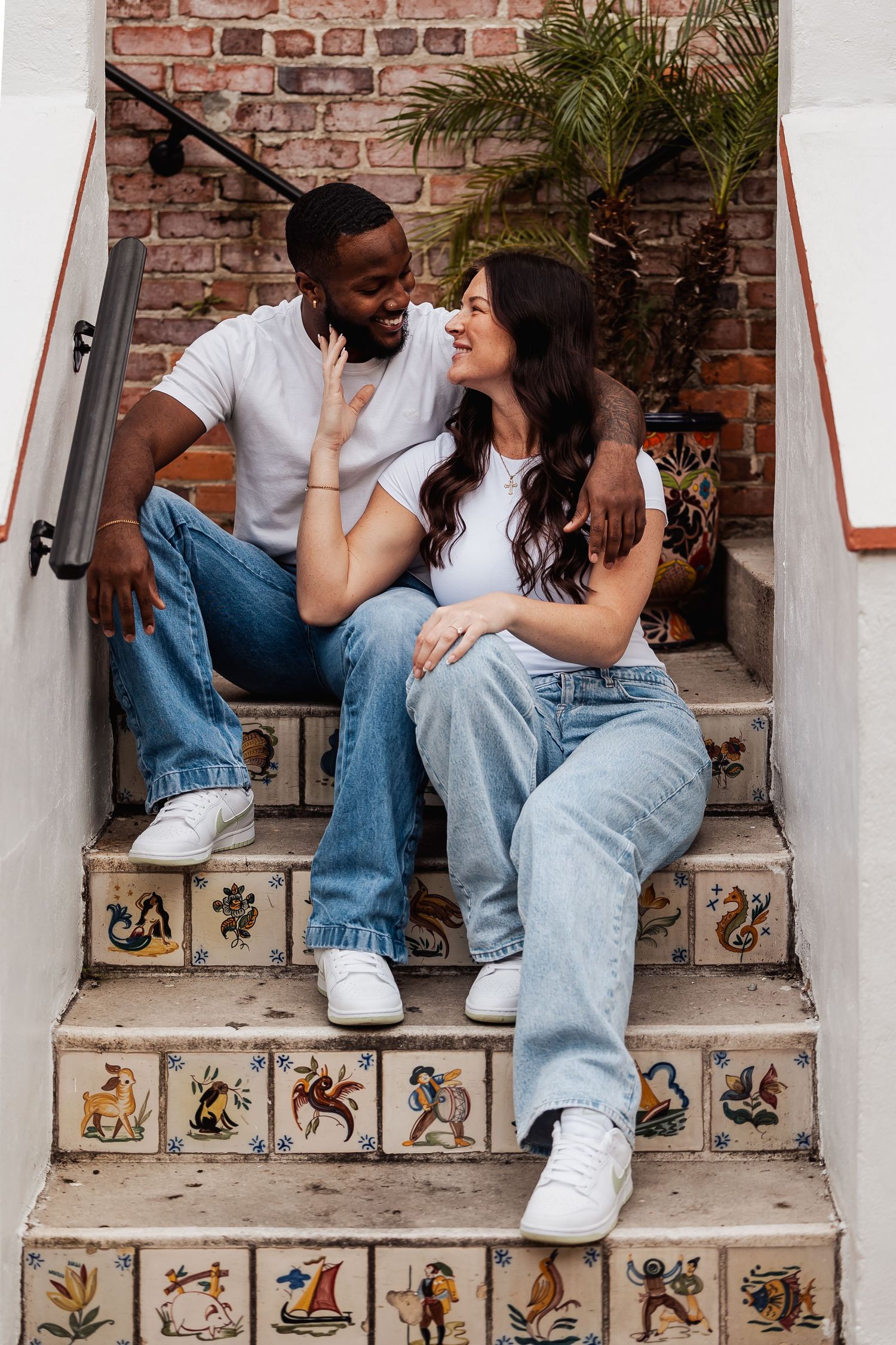 Happy interracial couple sitting close together on decorative tiled steps with brick wall backdrop.