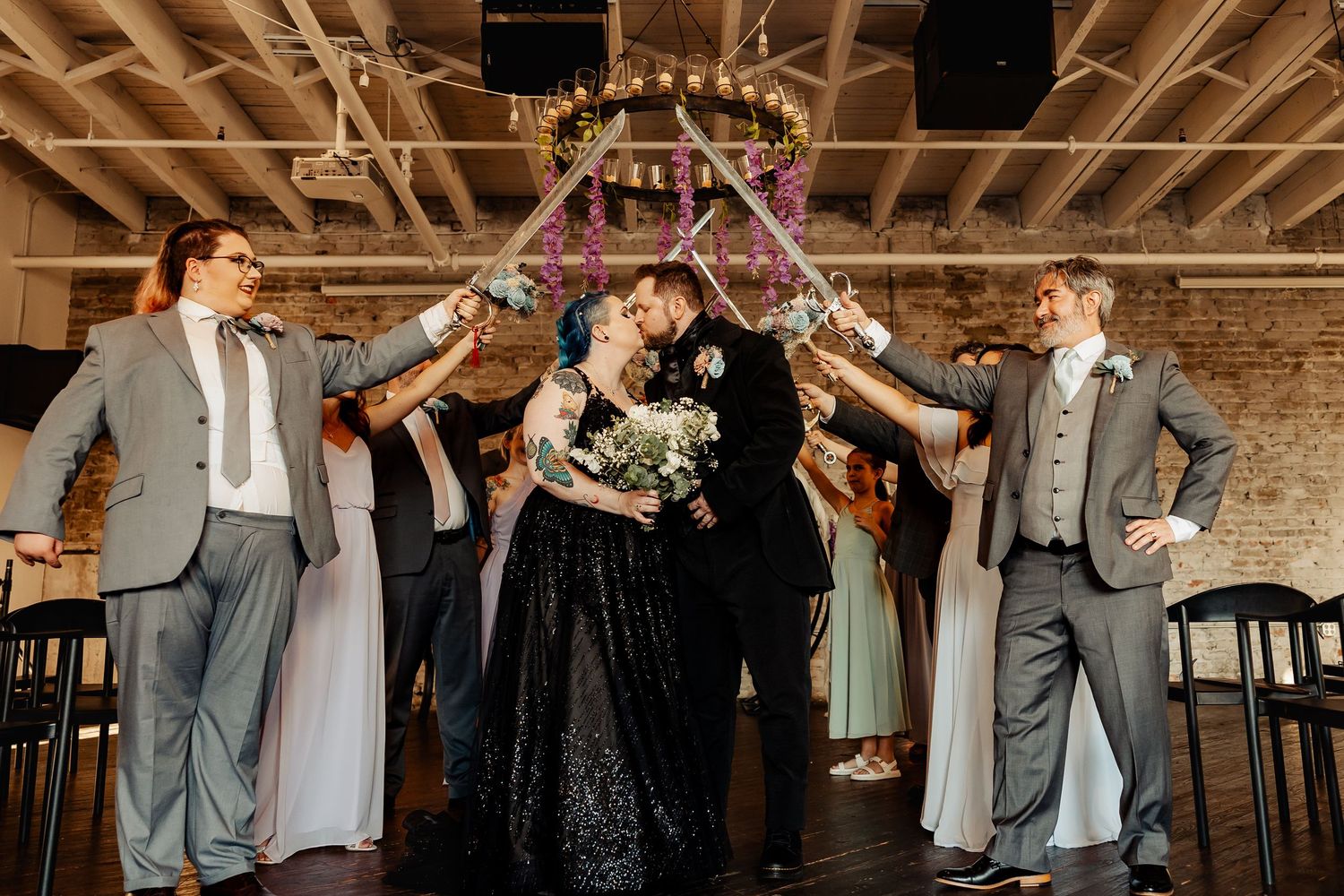Bride and groom share a kiss under raised swords held by wedding party in industrial venue.