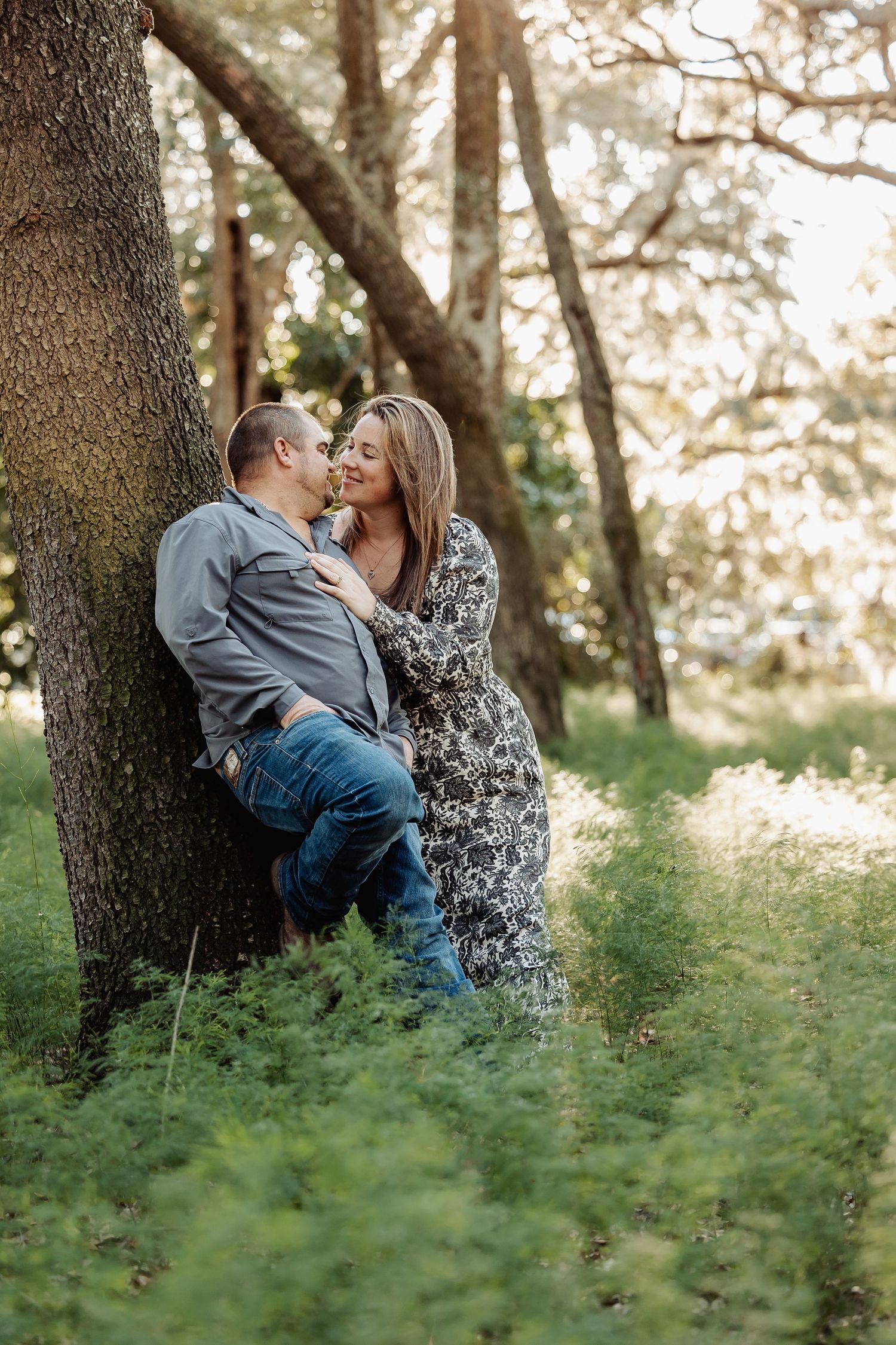 A couple shares a romantic kiss while leaning against a large tree in a sunlit forest.