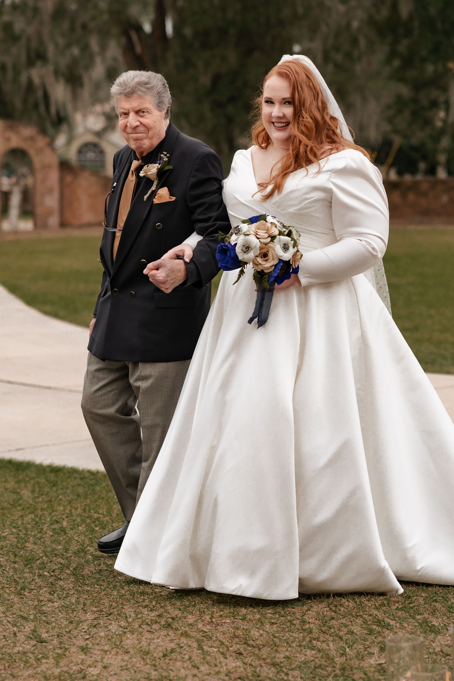Bride in white gown holding bouquet walks with older man in black suit on garden path.