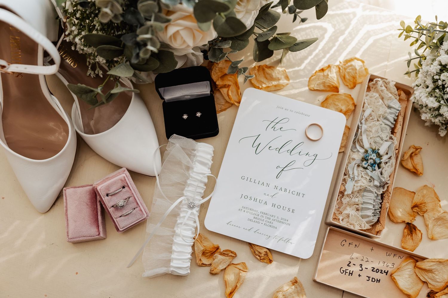 Wedding details flat lay with white shoes, rings, invitation and floral elements on beige background.
