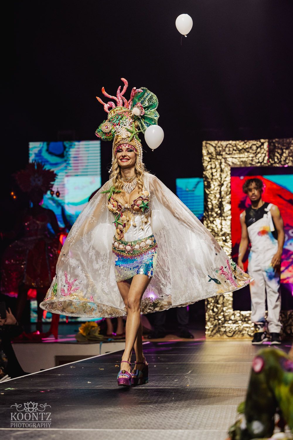 Fashion model walks runway in flowing white sheer cape and sparkly bodysuit during glamorous fashion show performance.