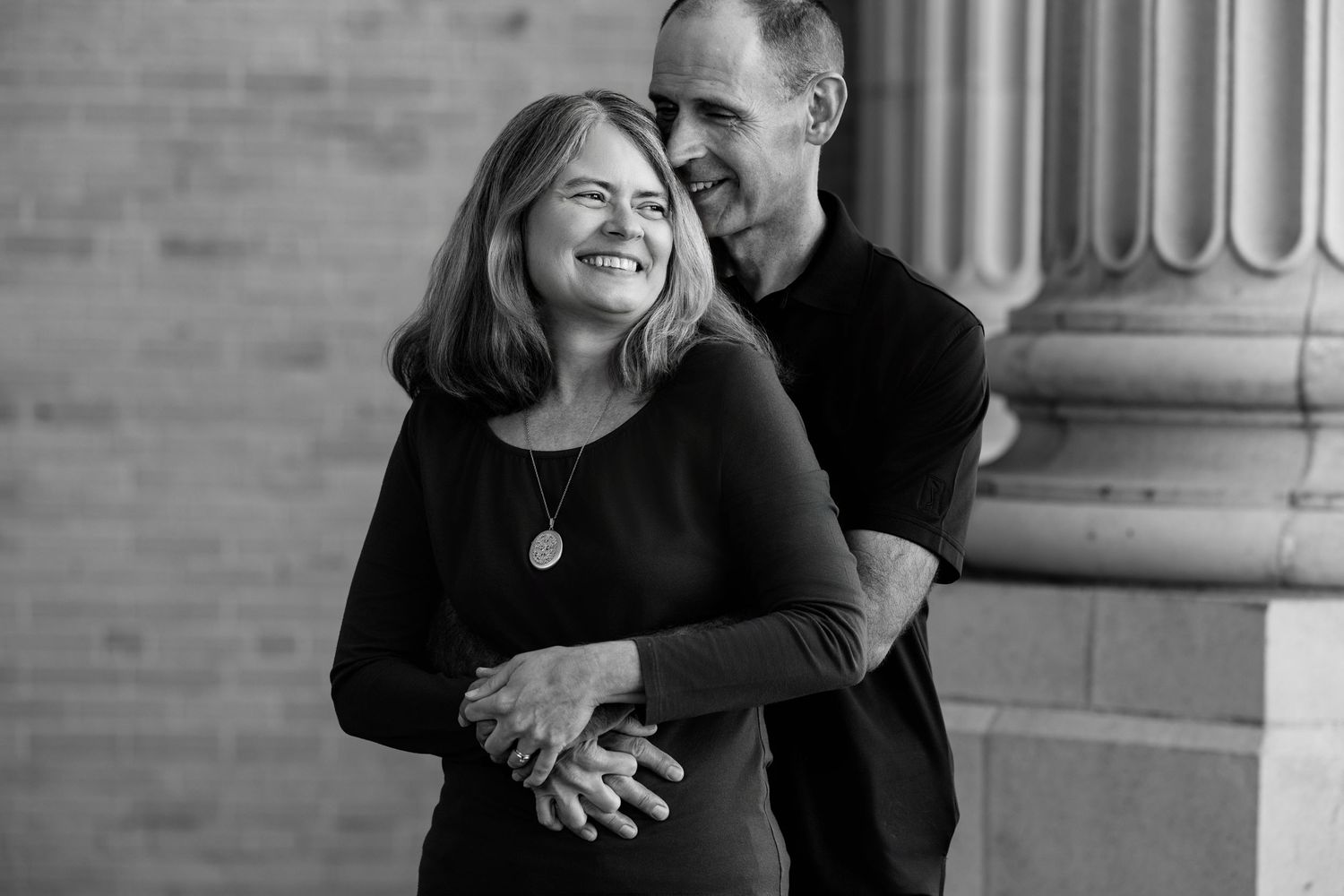 Black and white photo of couple embracing and laughing near stone columns outdoors.