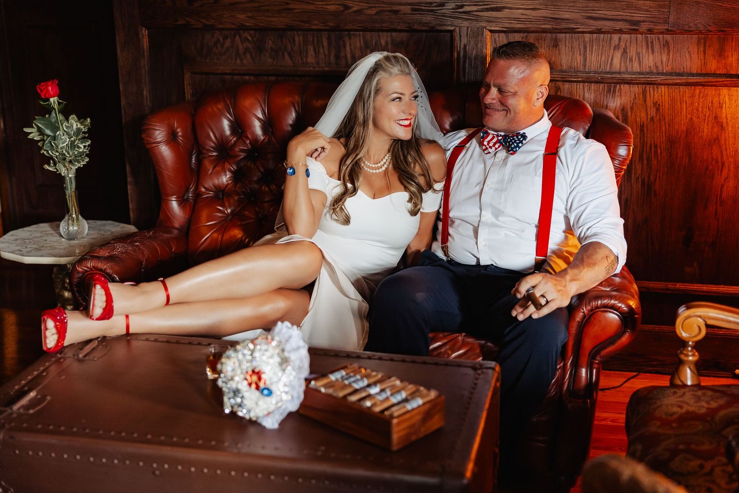 Bride and groom relaxing on leather sofa with cigars and bouquet, bride wearing veil and red heels.