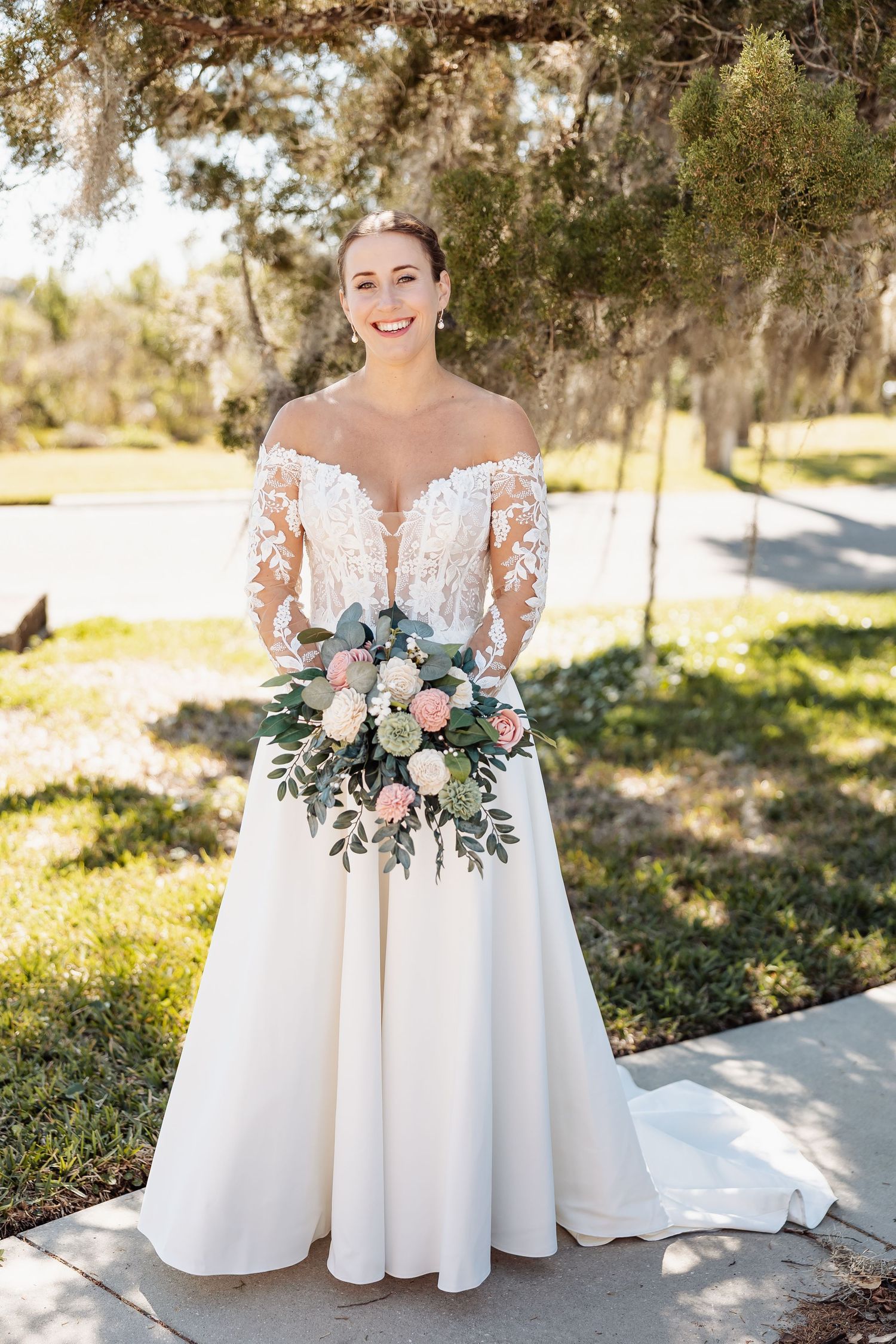 Bride in off-shoulder lace wedding gown holding a pink and green bouquet outdoors.