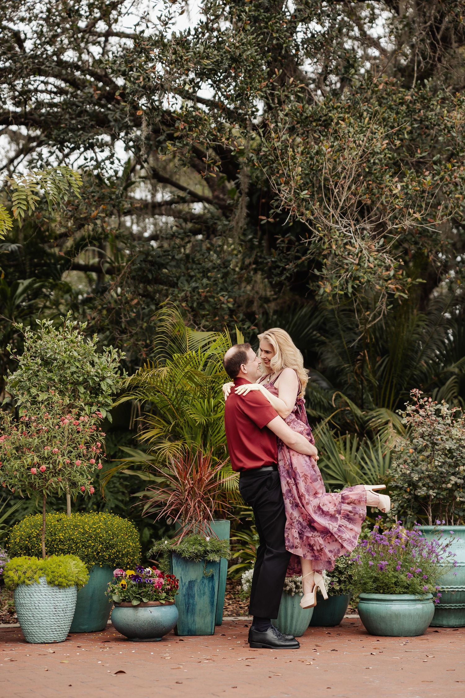 Romantic couple embracing and lifting among colorful potted plants in a lush garden setting.