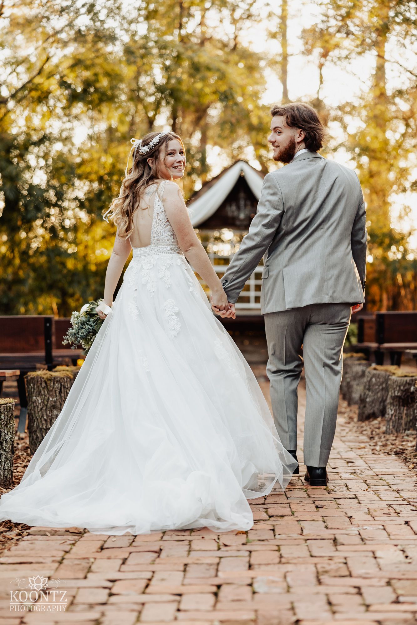 Bride and groom walk hand in hand along brick path toward rustic chapel at their outdoor woodland wedding.
