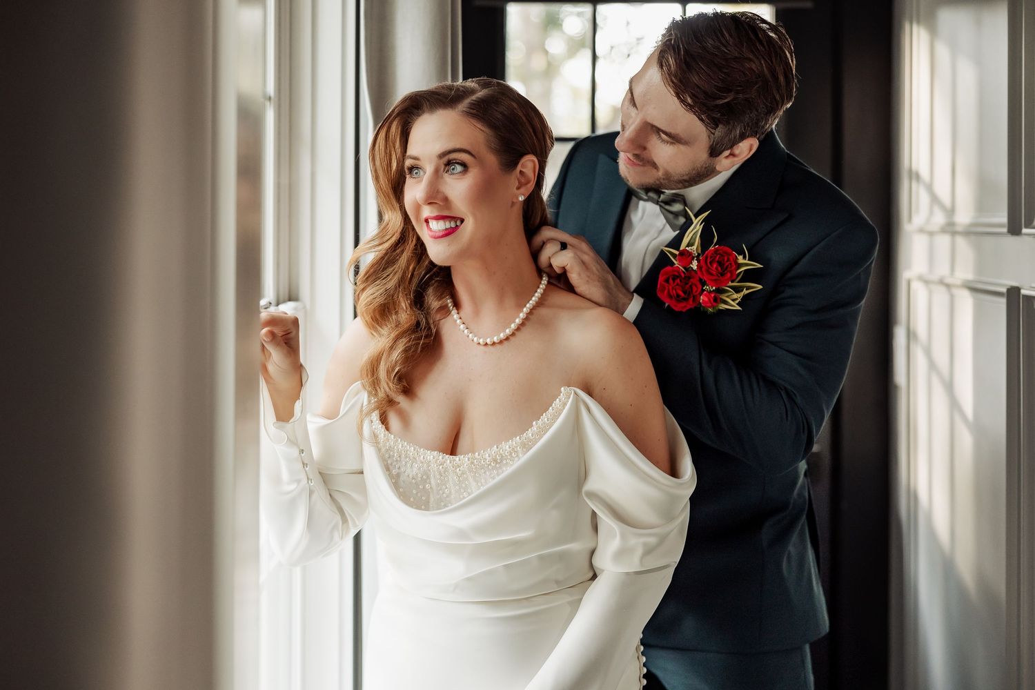 Bride in off-shoulder white gown smiling as groom adjusts her pearl necklace by window.