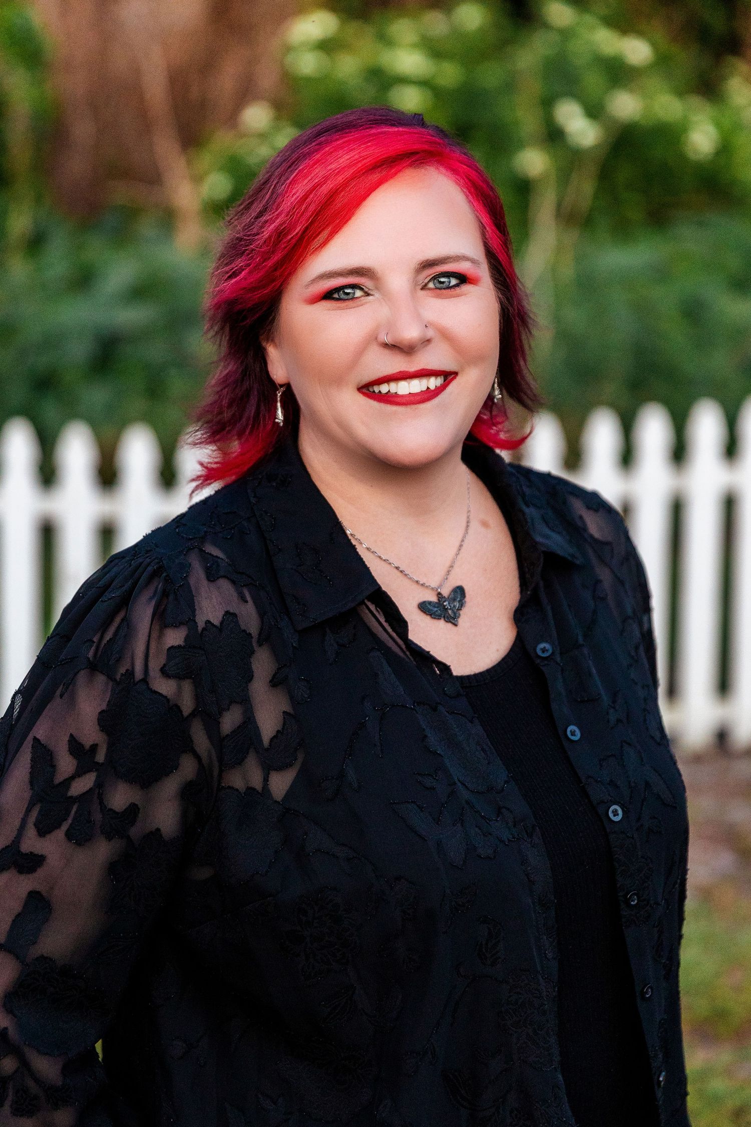 Smiling woman with bright red hair wearing black lace jacket stands before white picket fence outdoors.
