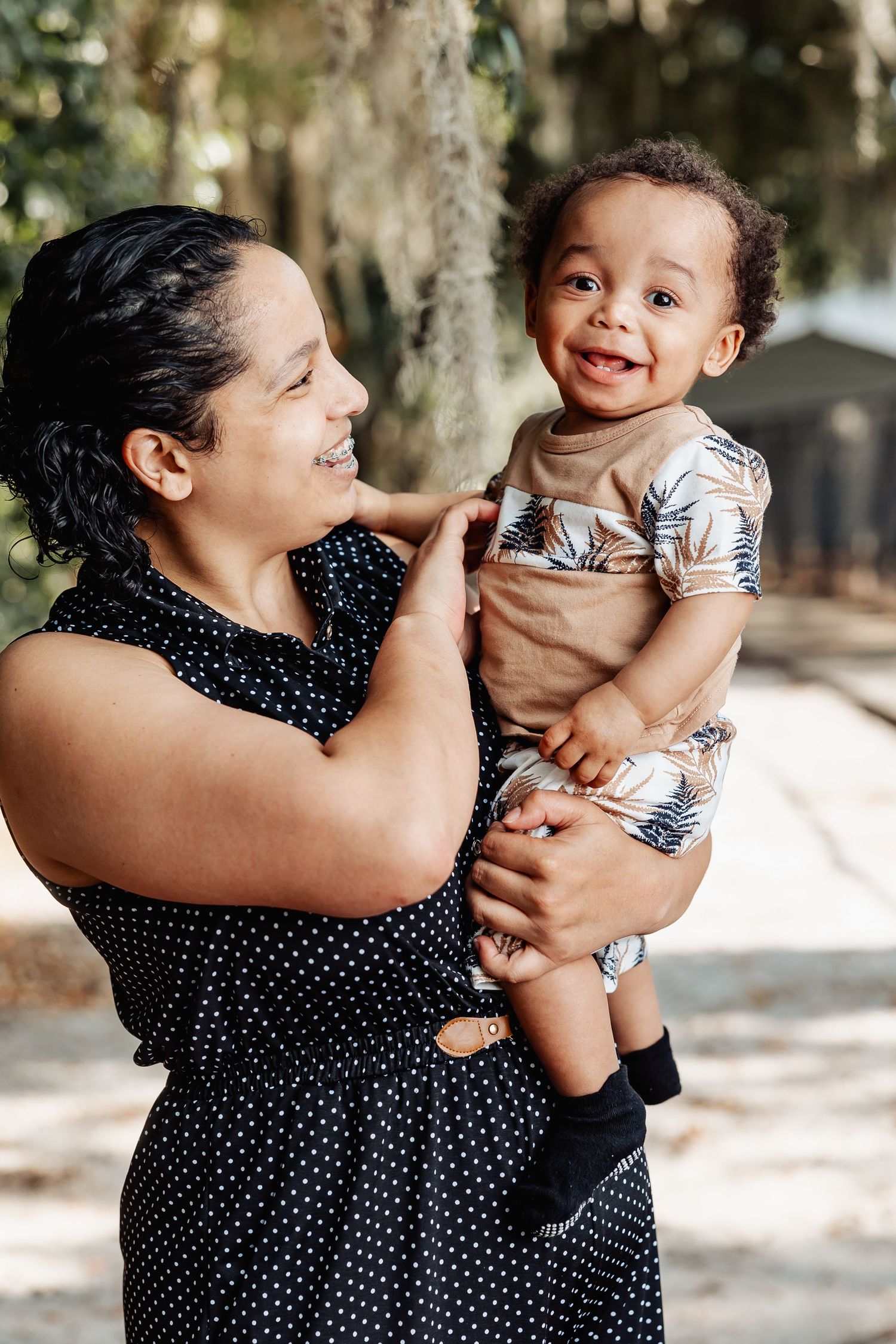 Smiling Hispanic mother holding cheerful baby boy in floral outfit in sunny outdoor park.