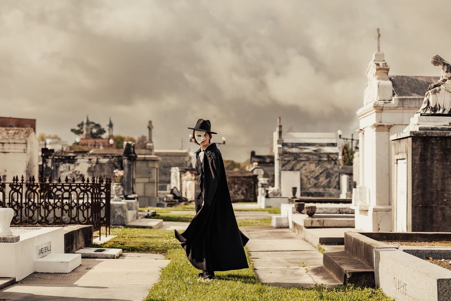 Elegant figure in flowing black cape and hat walking among white above-ground cemetery tombs.