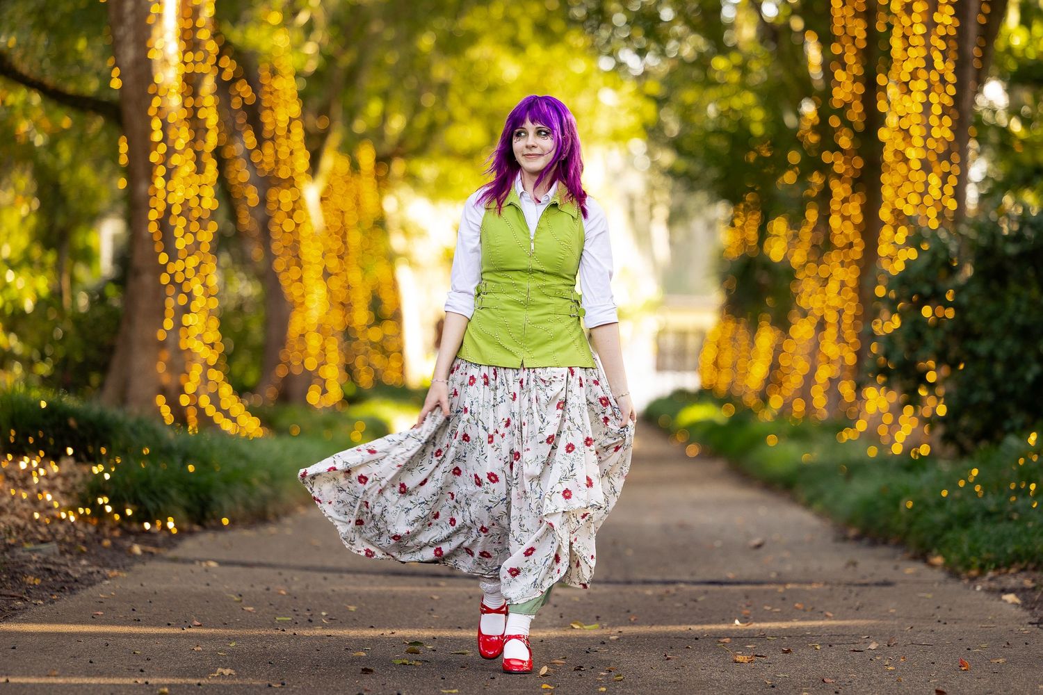 Woman with purple hair in green vest, floral skirt, and red shoes poses on a tree-lined path in golden light.
