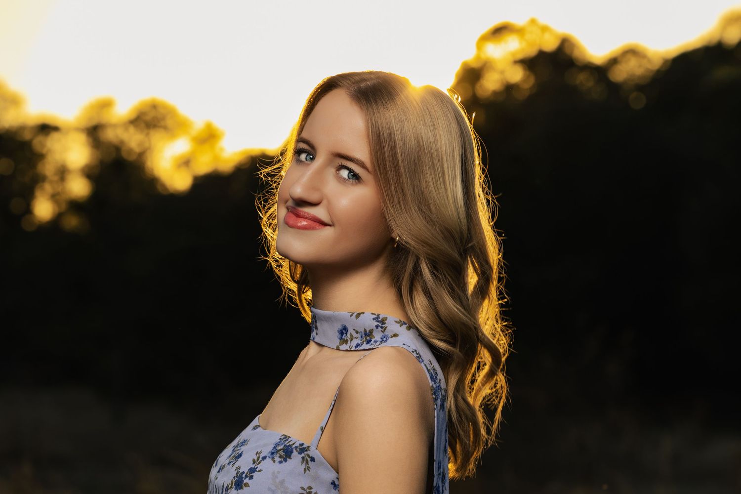 Young woman with wavy blonde hair in a floral dress smiling over her shoulder during golden hour sunset outdoors.