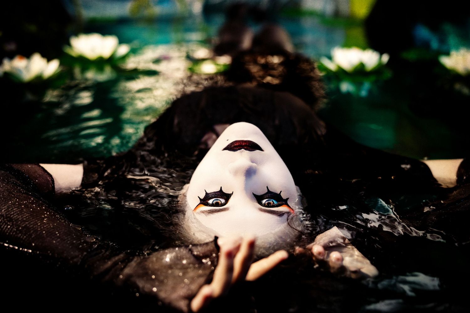 Woman wearing white theatrical mask floating in dark water with lily pads around her.