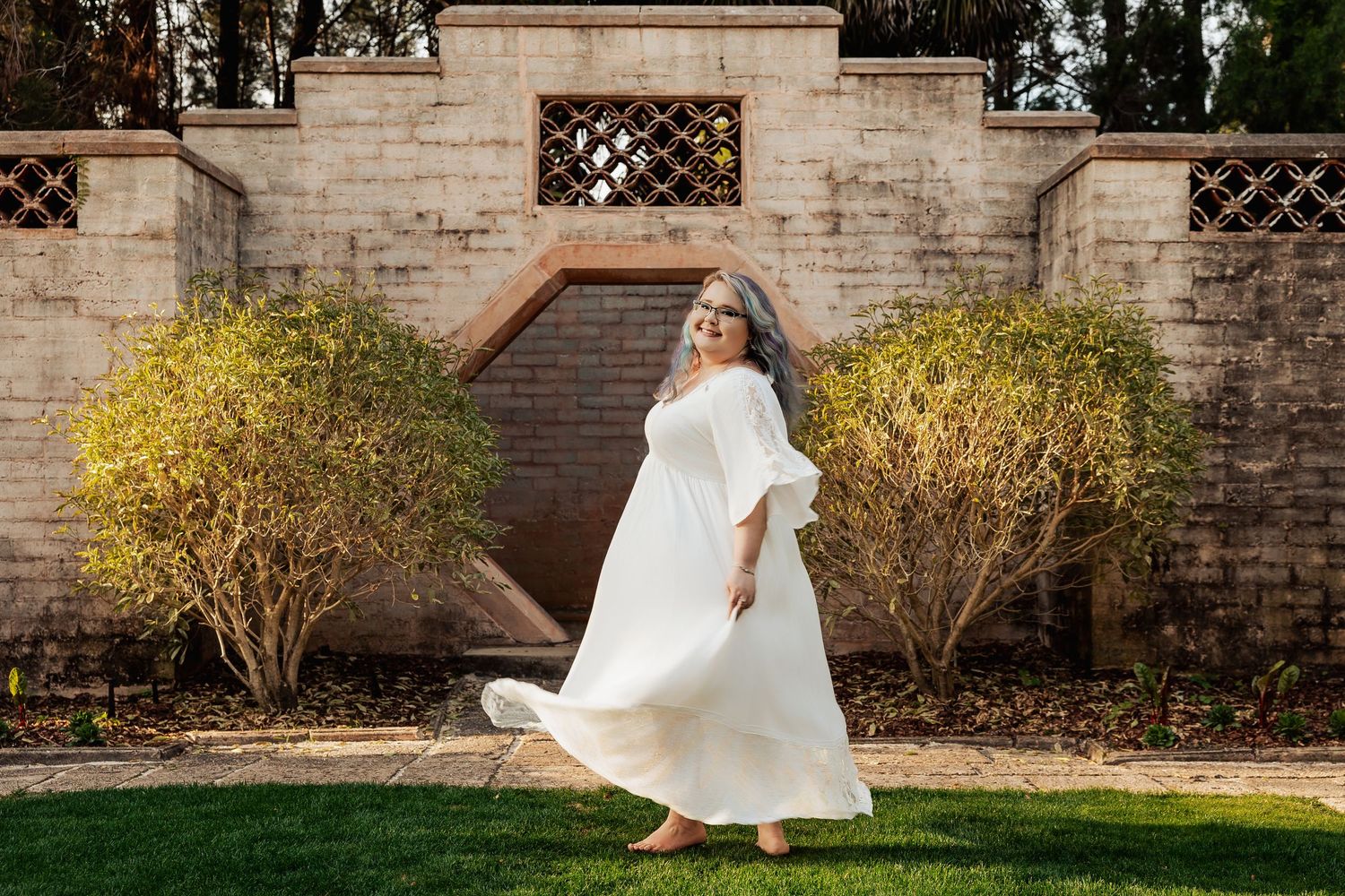 Woman in white flowing dress twirling in garden with stone wall backdrop.