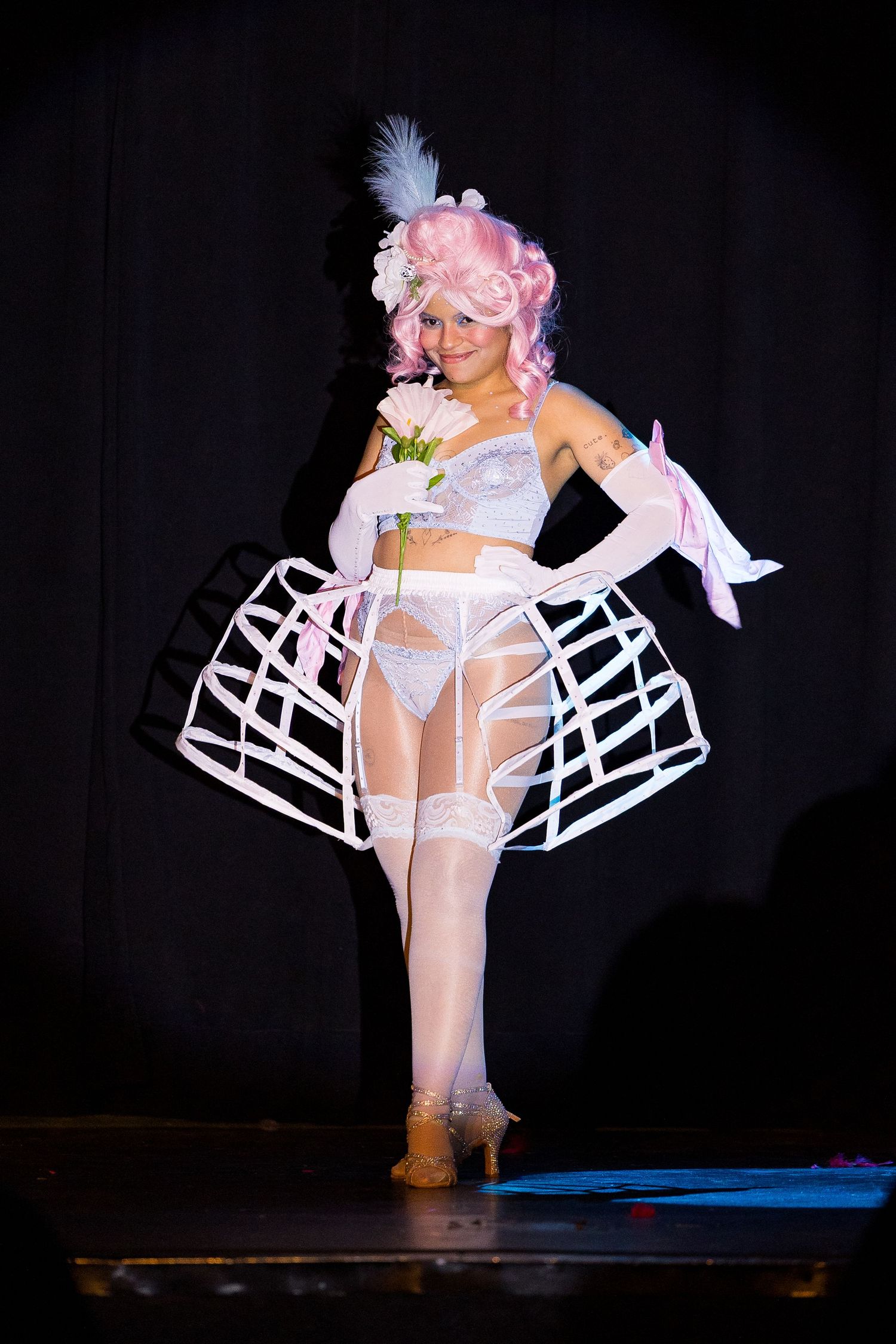 Burlesque performer in white lingerie with hoop skirt and pink wig holding flowers.