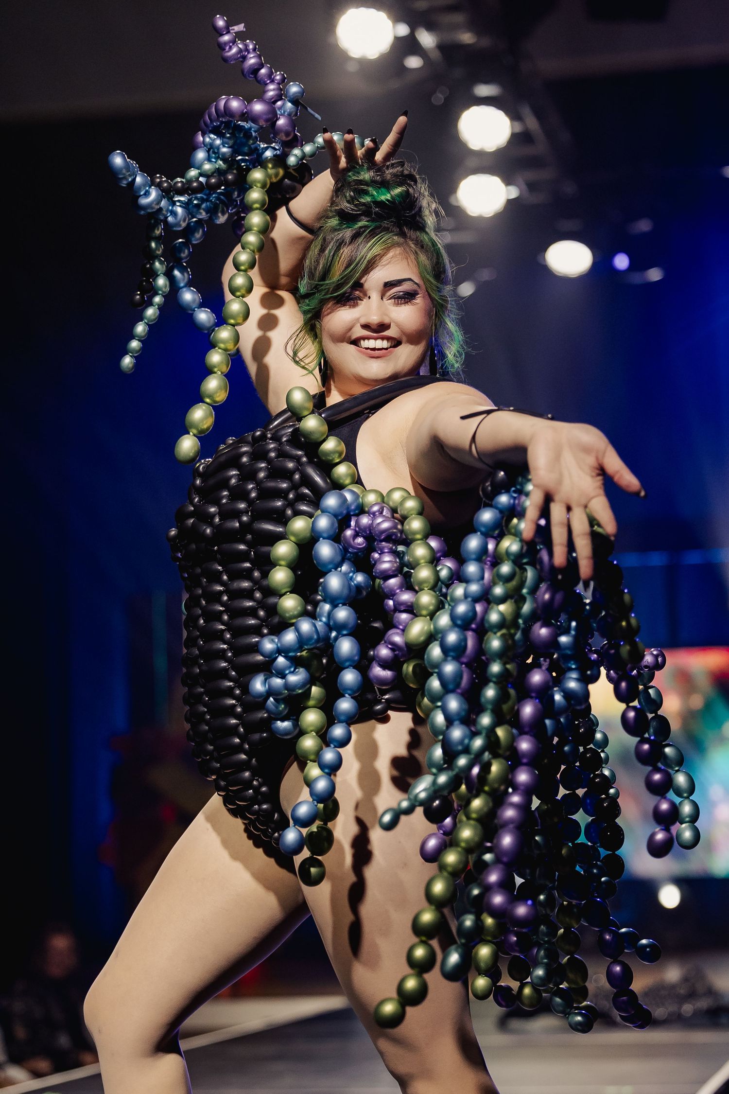 Model poses in black balloon bodysuit with colorful dangling balloon strands and matching headpiece.