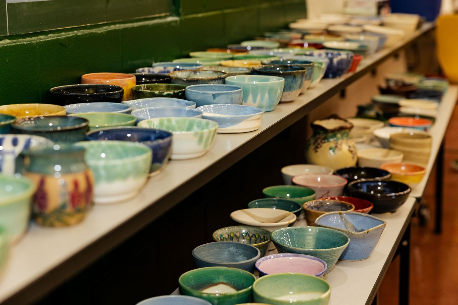 Rows of vibrant handmade ceramic bowls displayed on tables at an indoor charity event.