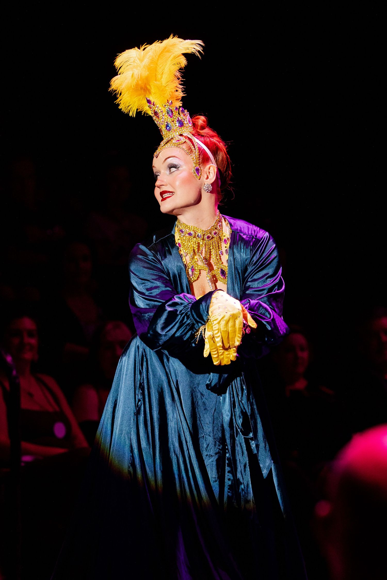 Performer in blue satin gown with yellow feather headdress and gold gloves on stage at burlesque cabaret.