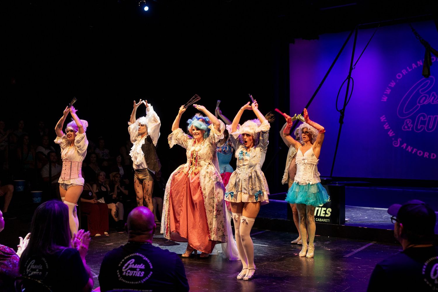 Burlesque performers in rococo-inspired costumes dance for front-row audience under purple stage lighting.