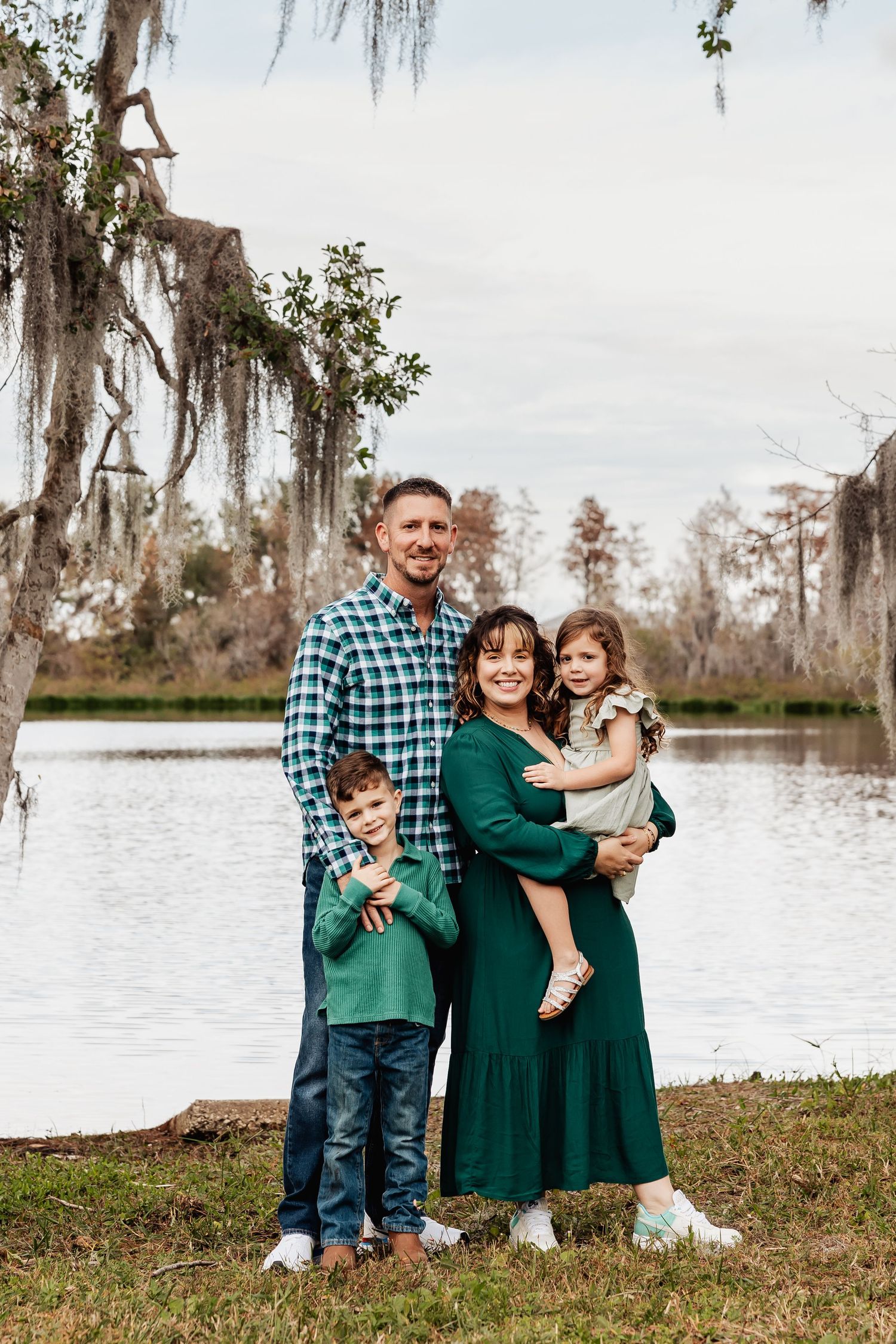 A smiling family of four poses together by a peaceful lake surrounded by Spanish moss-draped trees.