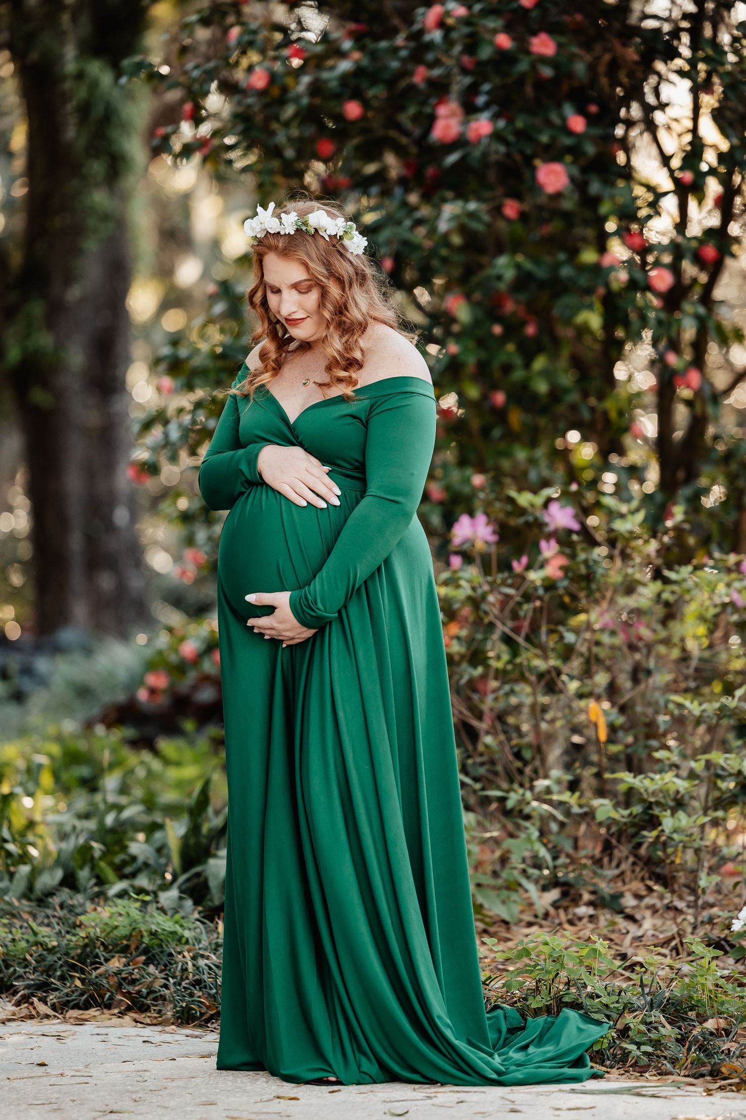Pregnant woman in flowing green off-shoulder gown with floral crown in garden setting.