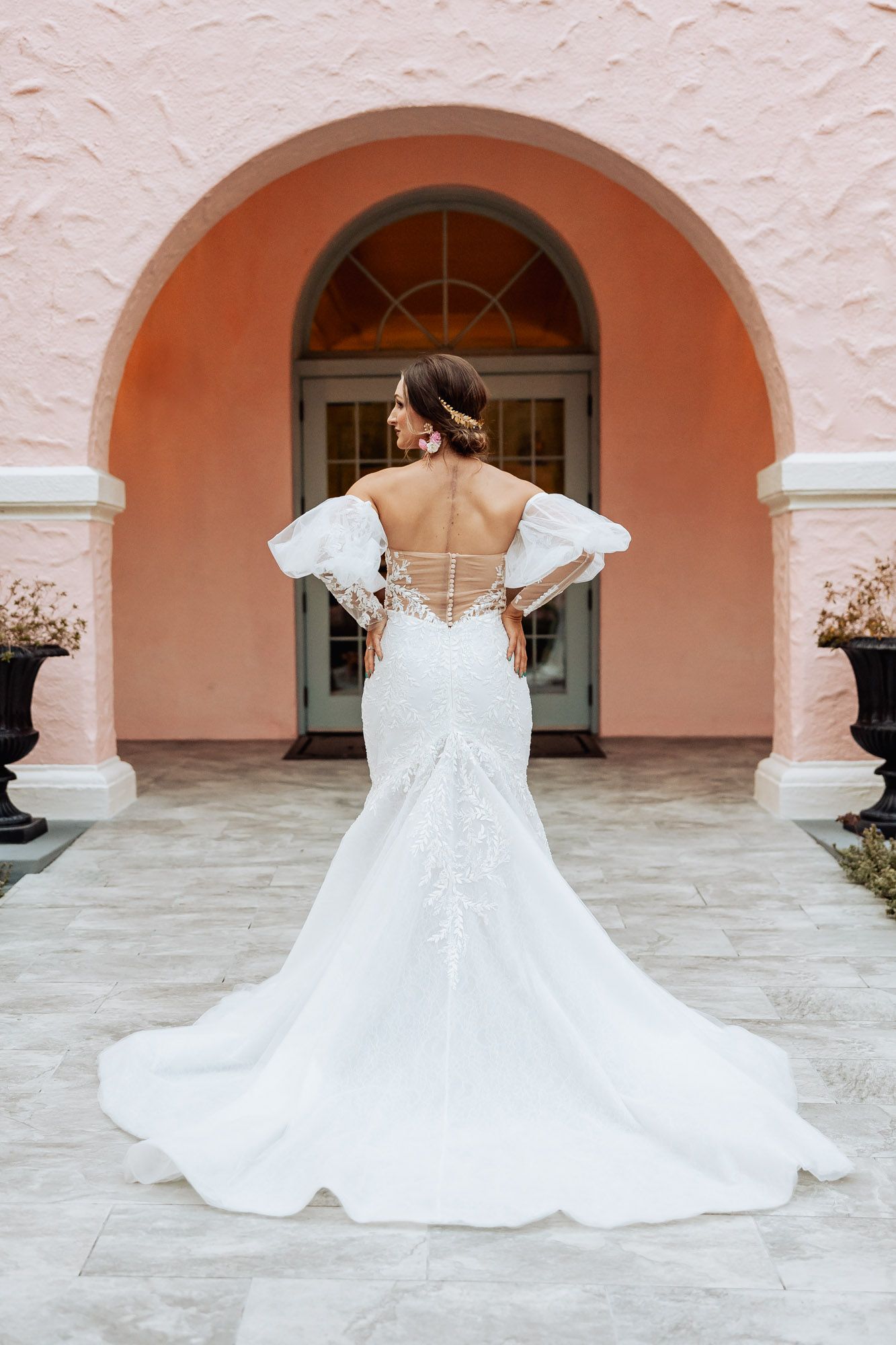 A bride wearing an elegant white mermaid wedding gown with puff sleeves stands in front of a pink arched entryway.