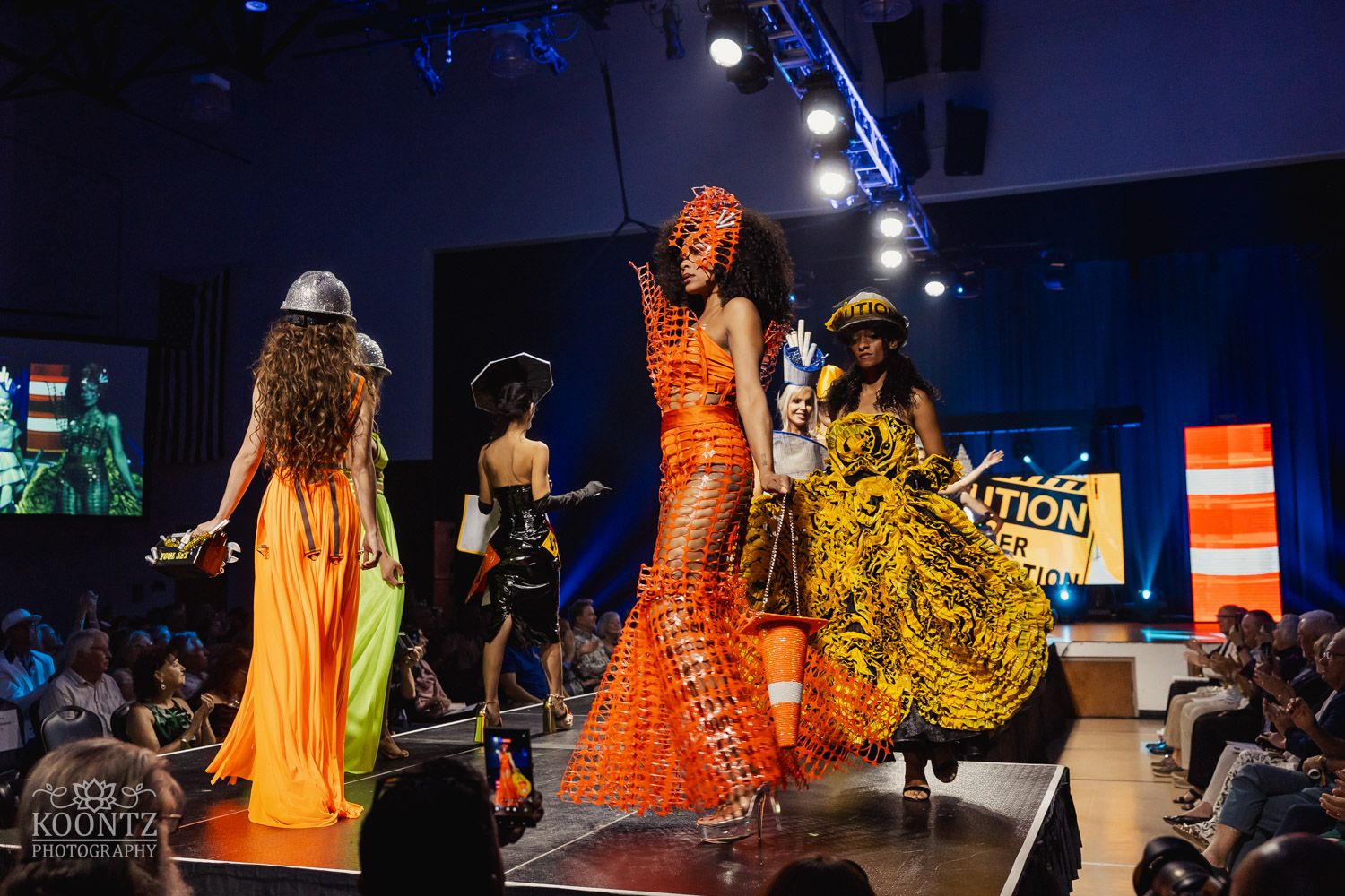 Fashion show featuring colorful orange dresses and theatrical costumes on a dark stage with dramatic lighting.