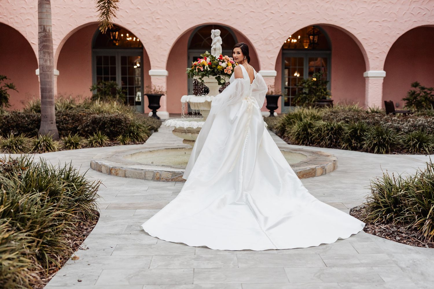 Bride in classic white ball gown with long train standing before pink arched venue fountain.
