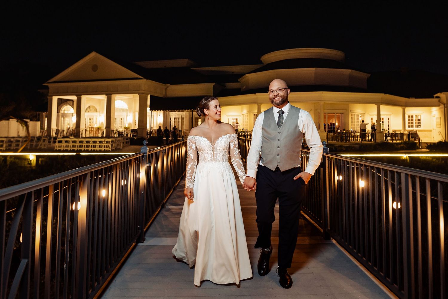 Bride and groom walking hand in hand on a lit bridge outside a wedding venue at night.