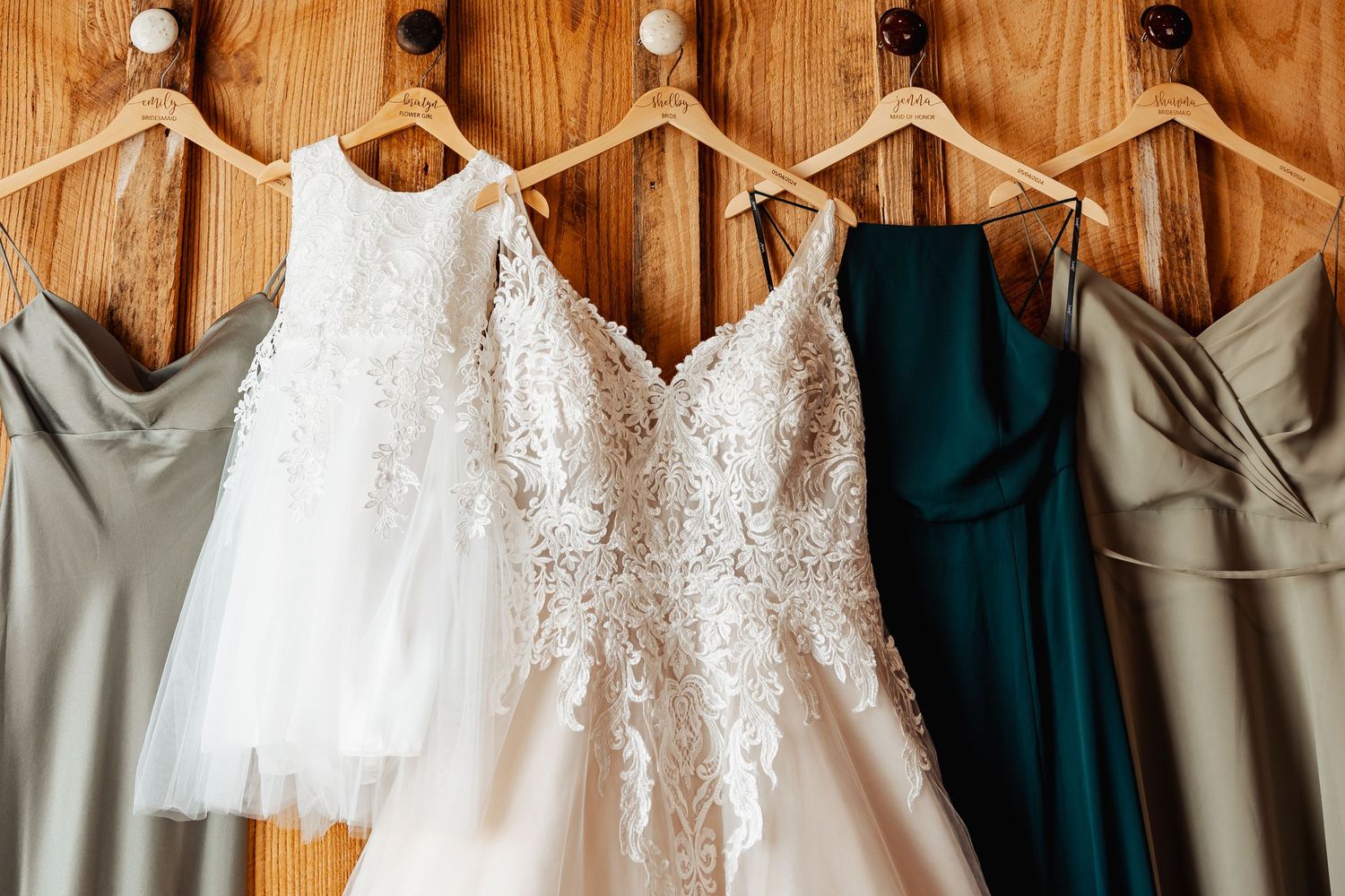 Wedding dress and bridesmaid gowns in white, gray and teal hanging on wooden wall hooks.