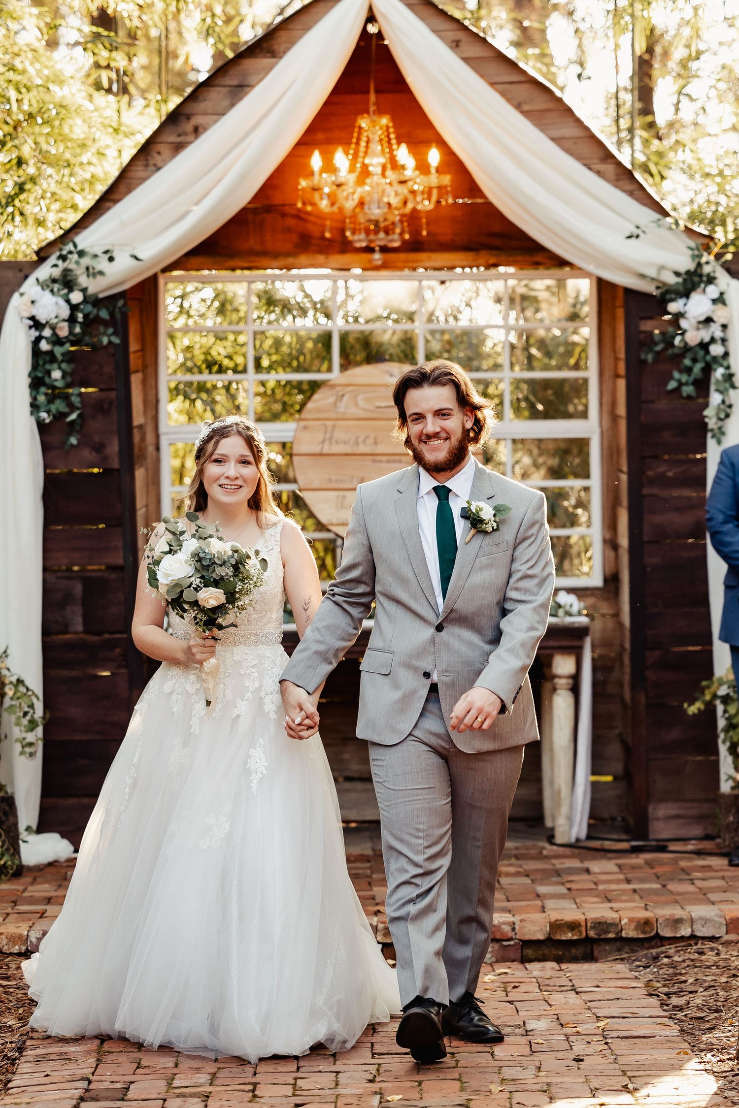 Newlyweds standing under wooden arch decorated with white draping and crystal chandelier.