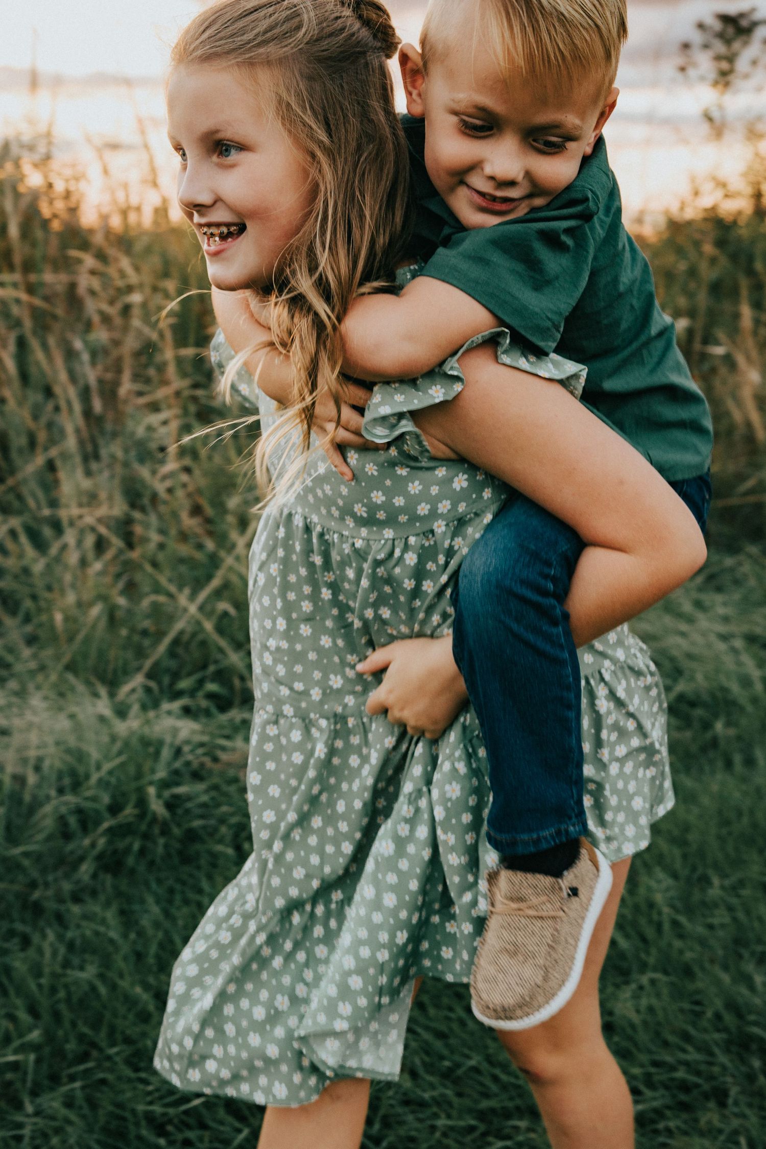 Young siblings playing and giving piggyback rides in a grassy field at sunset.