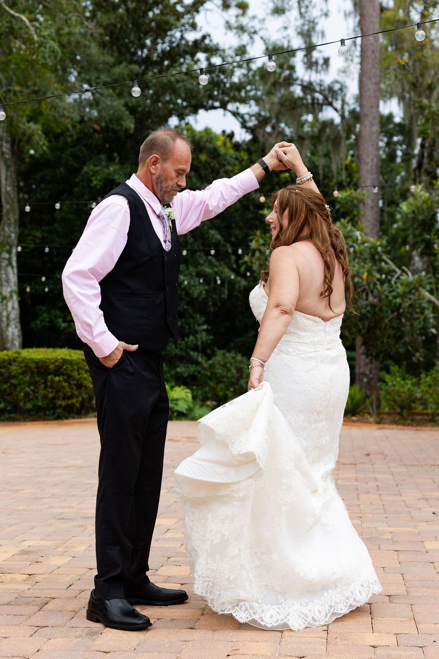 Groom twirls bride in elegant lace wedding gown on brick patio under romantic string lights outdoors.